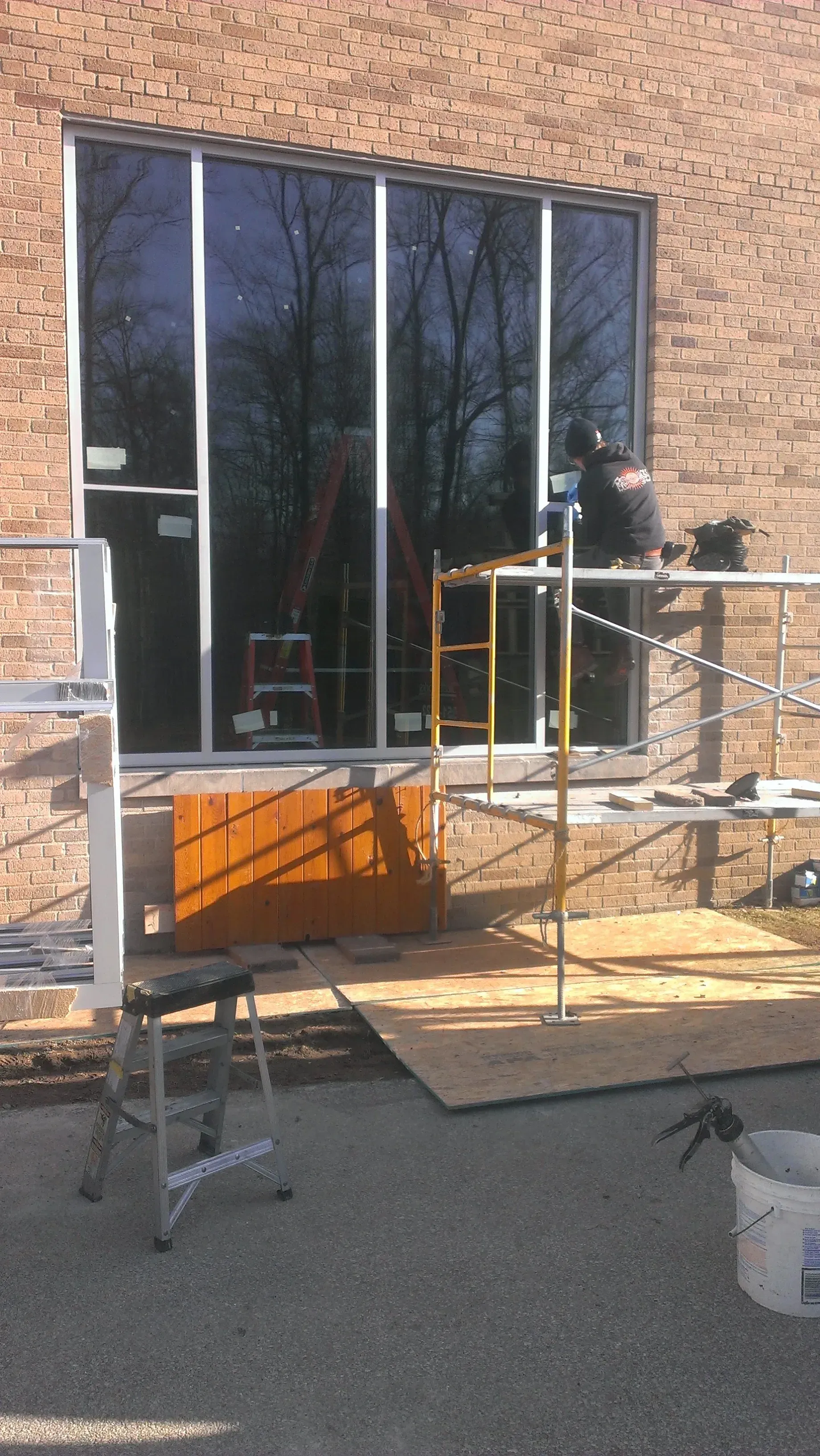 Construction workers installing a large window in a brick building. Scaffolding and a ladder are present.