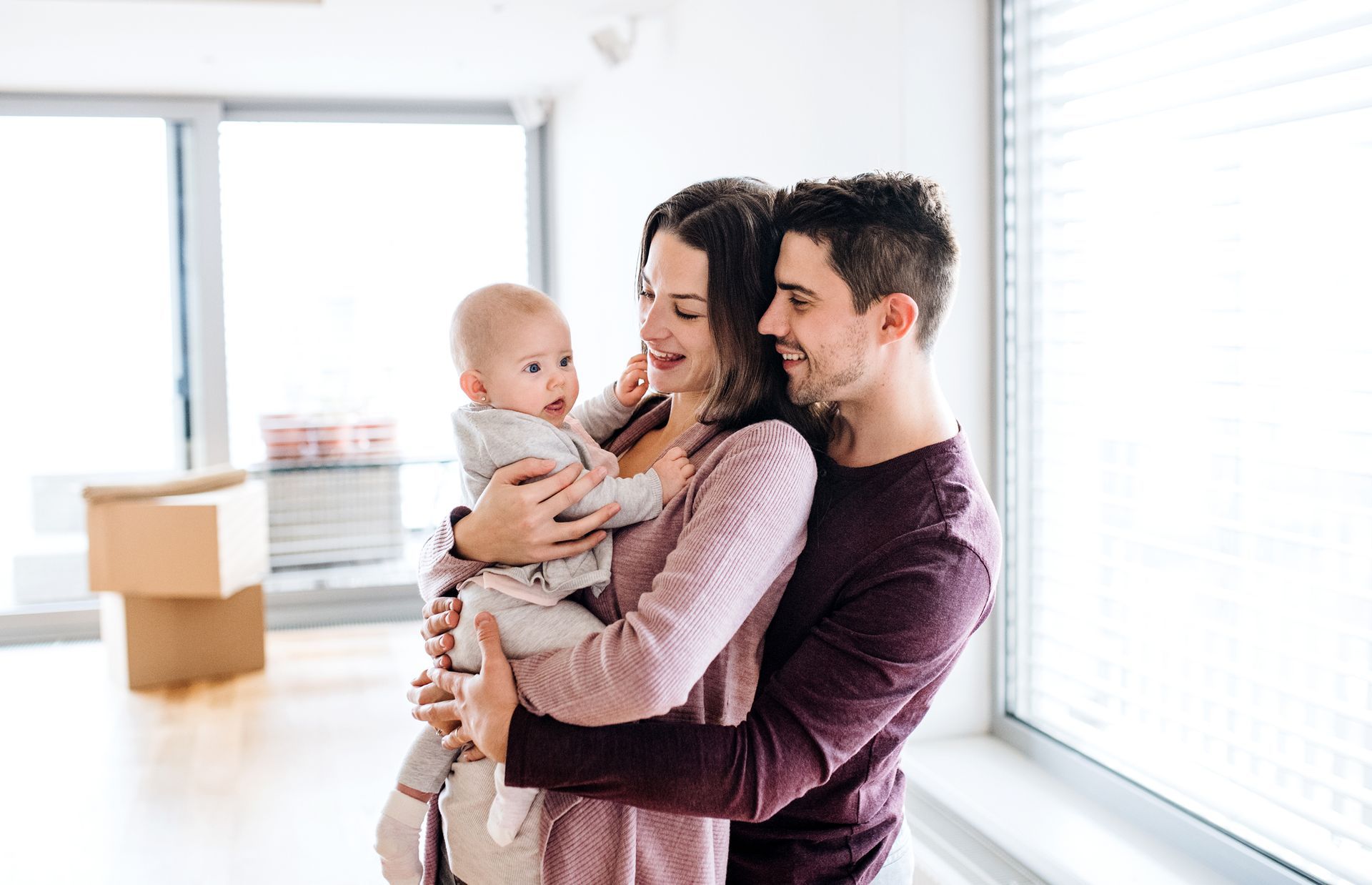 Parents holding a baby near a window, boxes in the background. They are smiling.