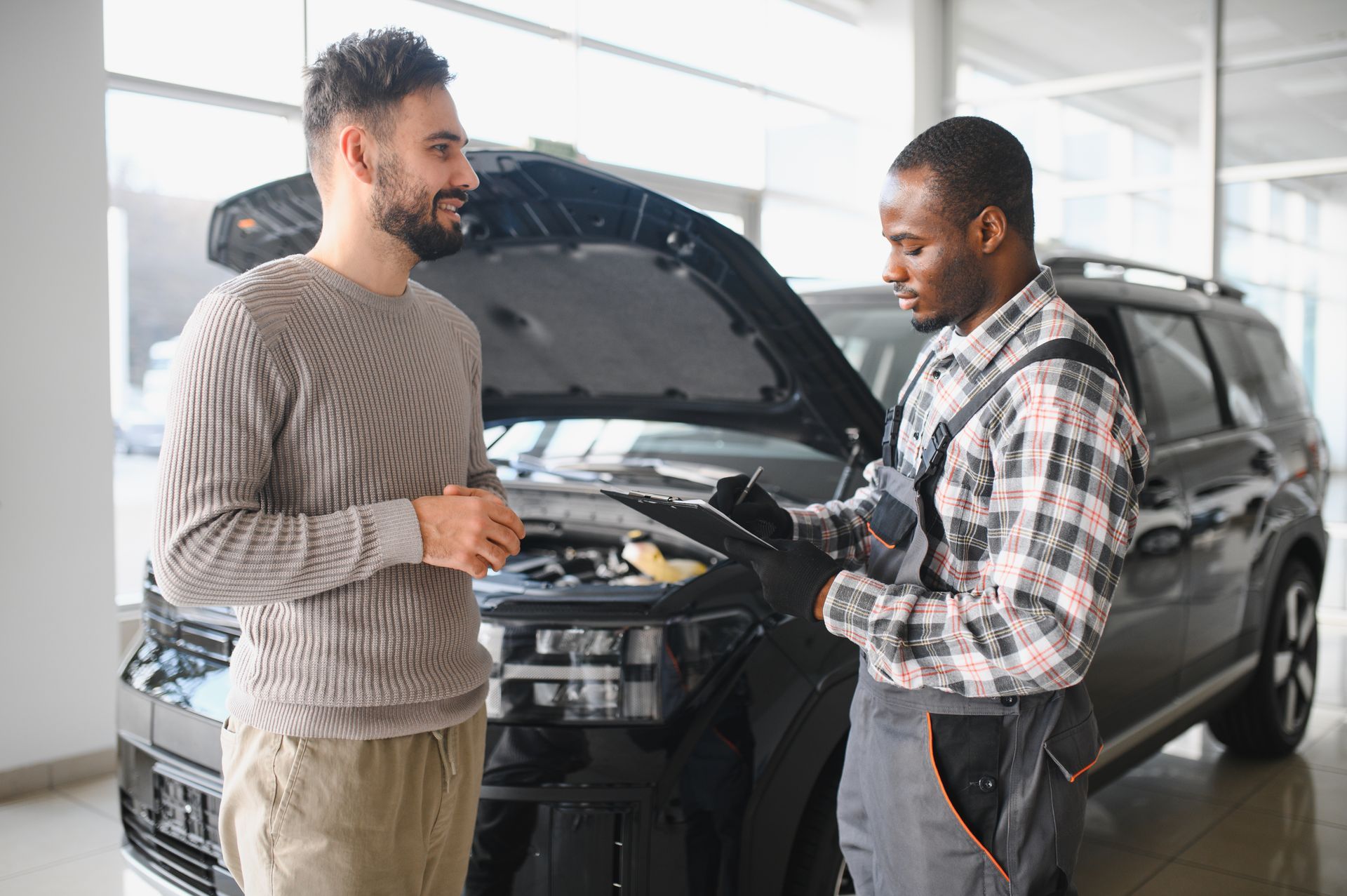 Mechanic with clipboard talking to a person next to a black car with the hood open in a service shop.