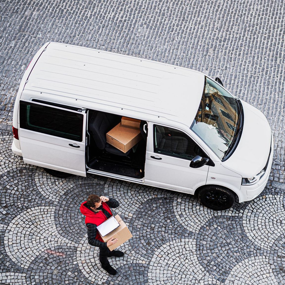White delivery van with open door; person in red jacket carrying packages. Cobblestone street.