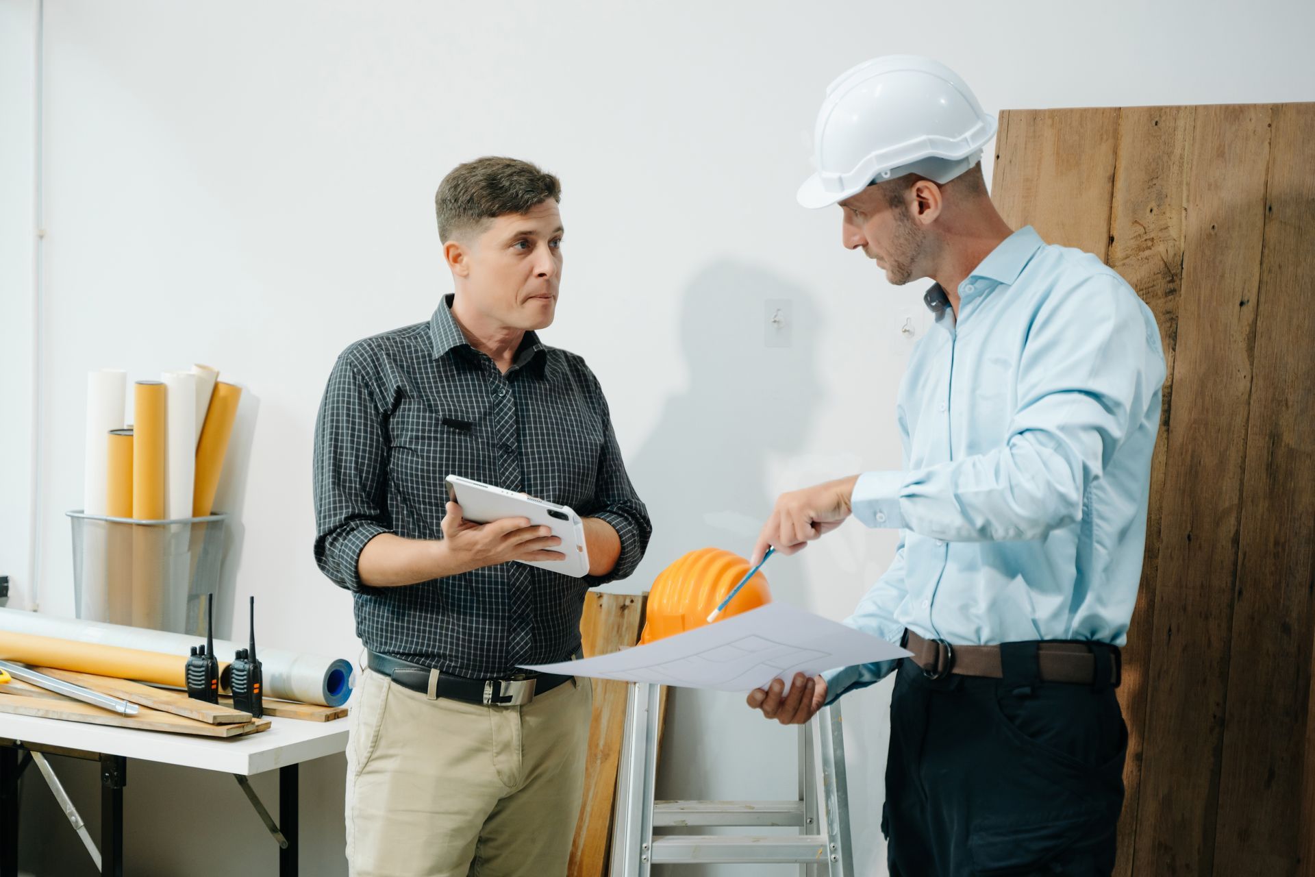 Two men in a construction setting reviewing blueprints; one wears a hard hat and points, the other holds a tablet.