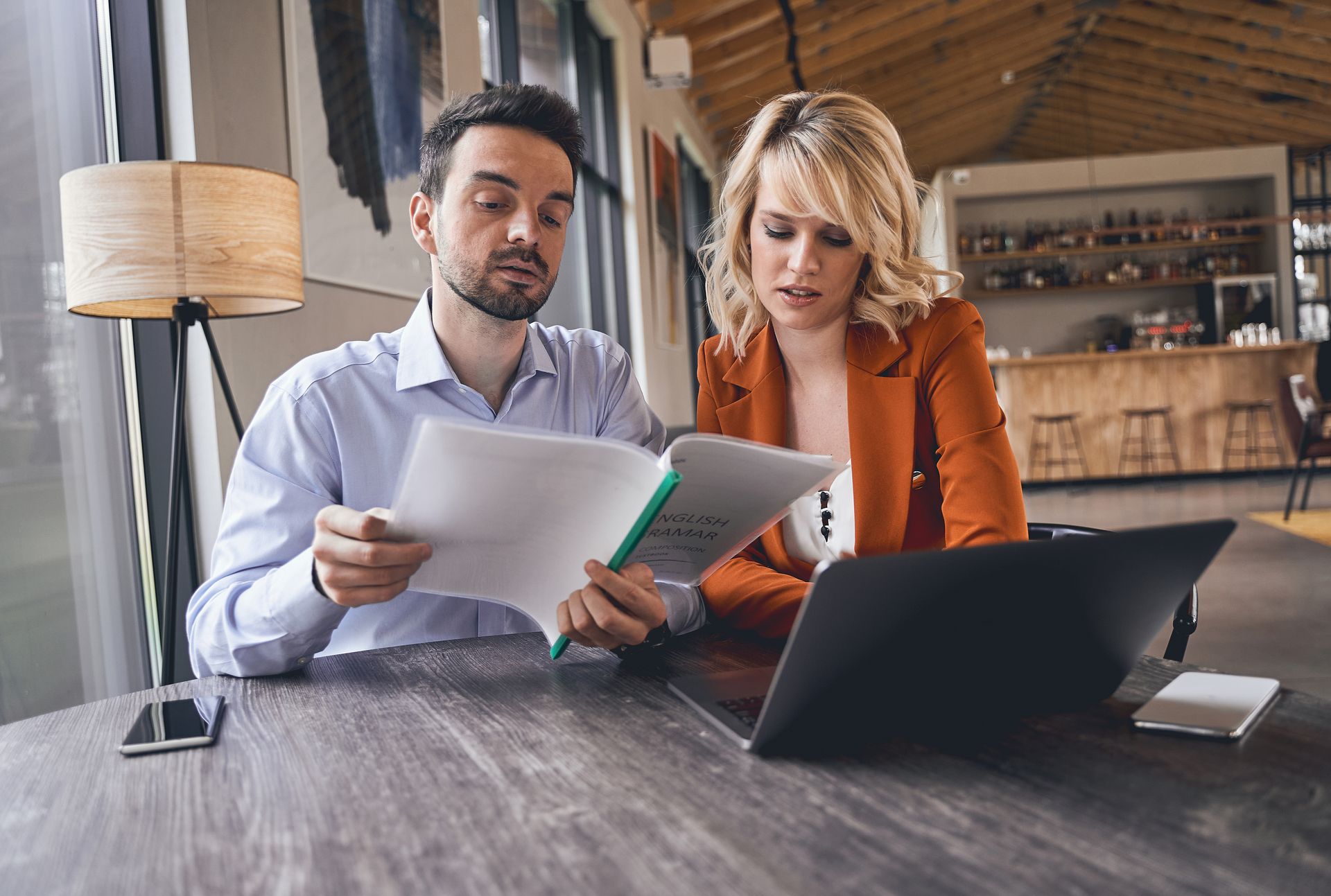 Man and woman reviewing documents together at a table with laptop and phone in a cafe setting.