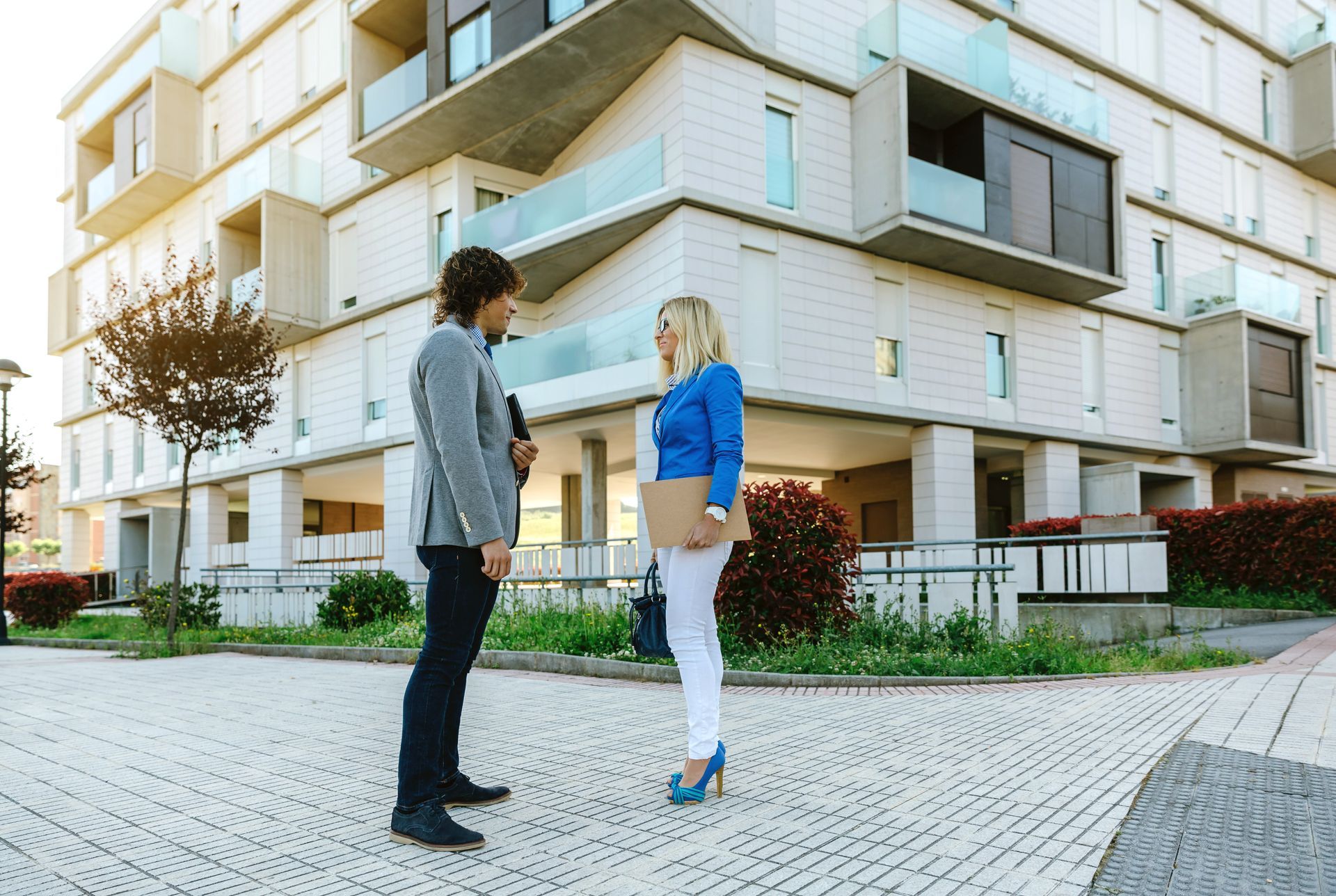 Two people in front of a modern building; woman holds a notepad, they're talking.