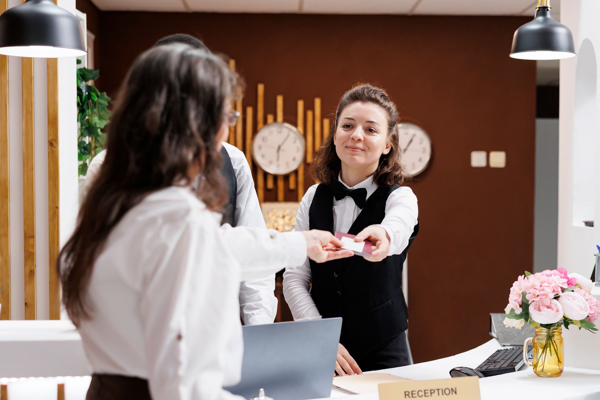 Hotel receptionist handing a card to a guest at the front desk. Brown wall, clocks, and flower vase in background.