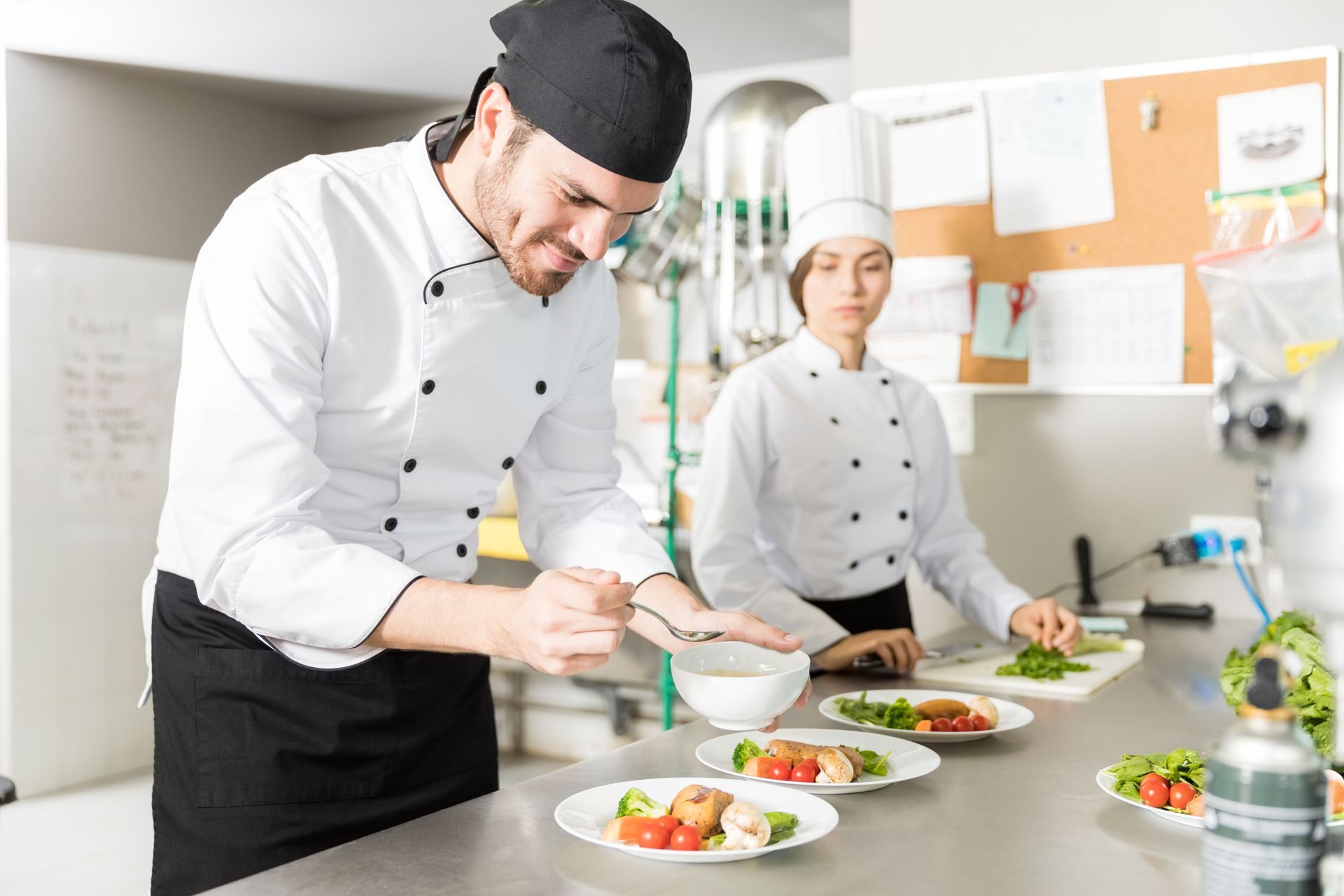 Two chefs in a commercial kitchen preparing plates. One garnishes a plate, the other preps vegetables.