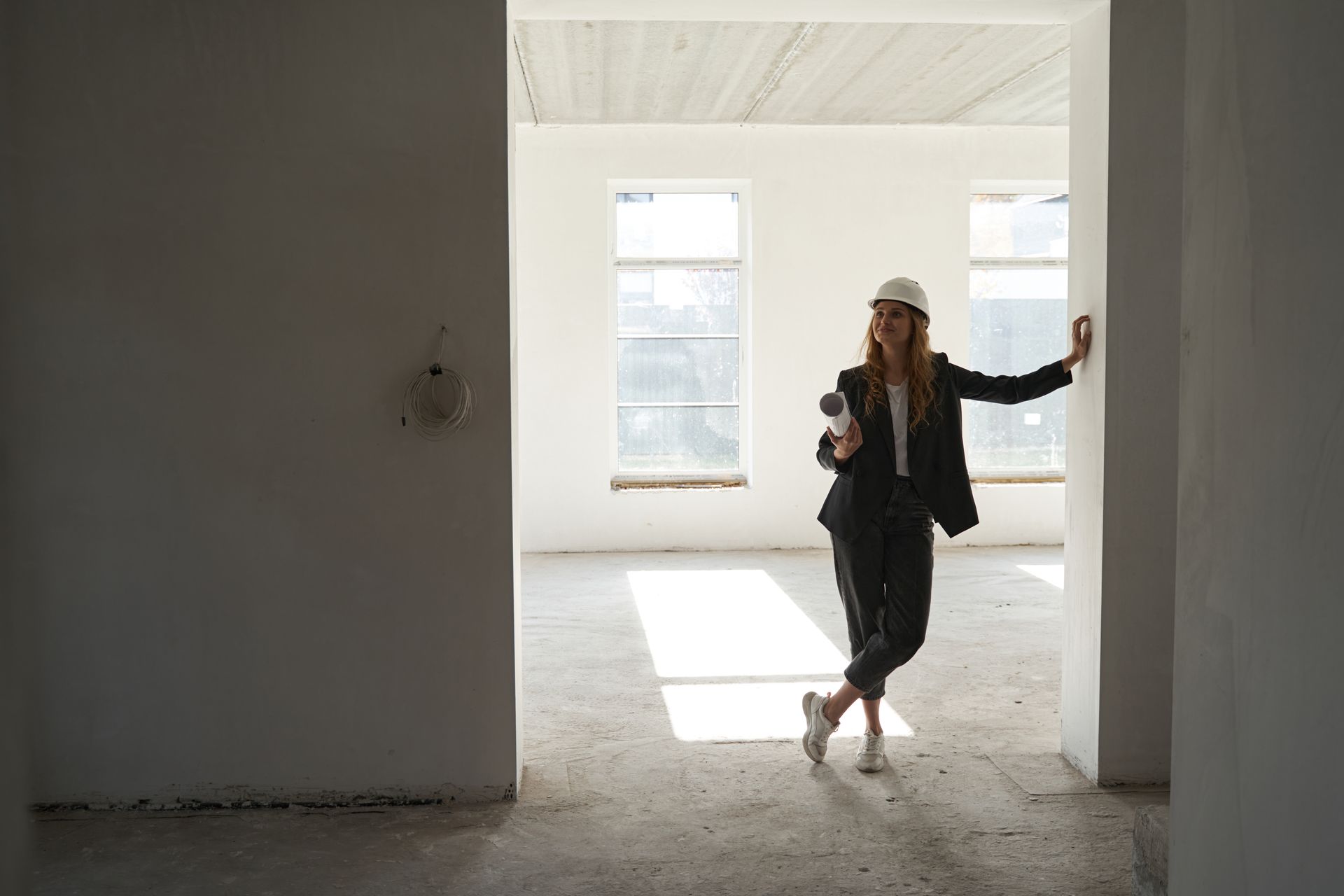 Woman in a hard hat and suit leans against a wall in an unfinished building.