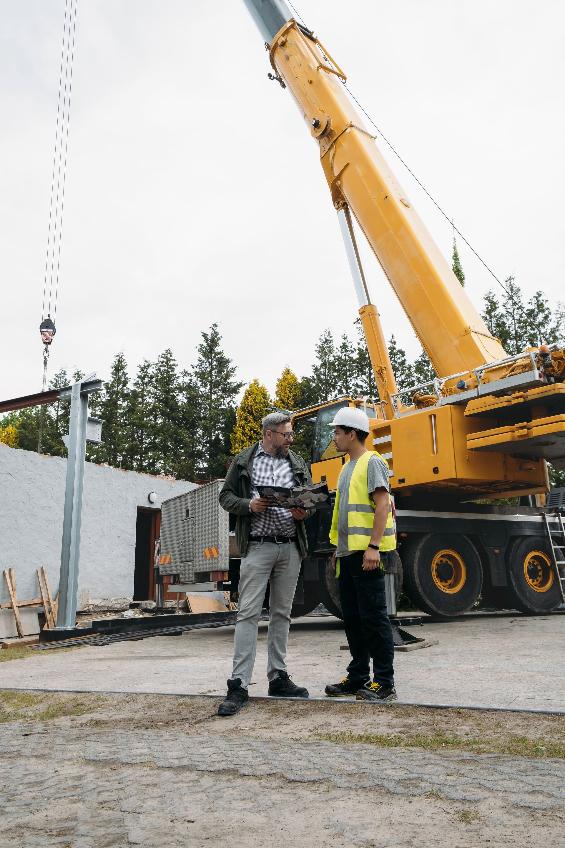 Two men discussing construction plans near a yellow crane. One wears a hard hat and safety vest.
