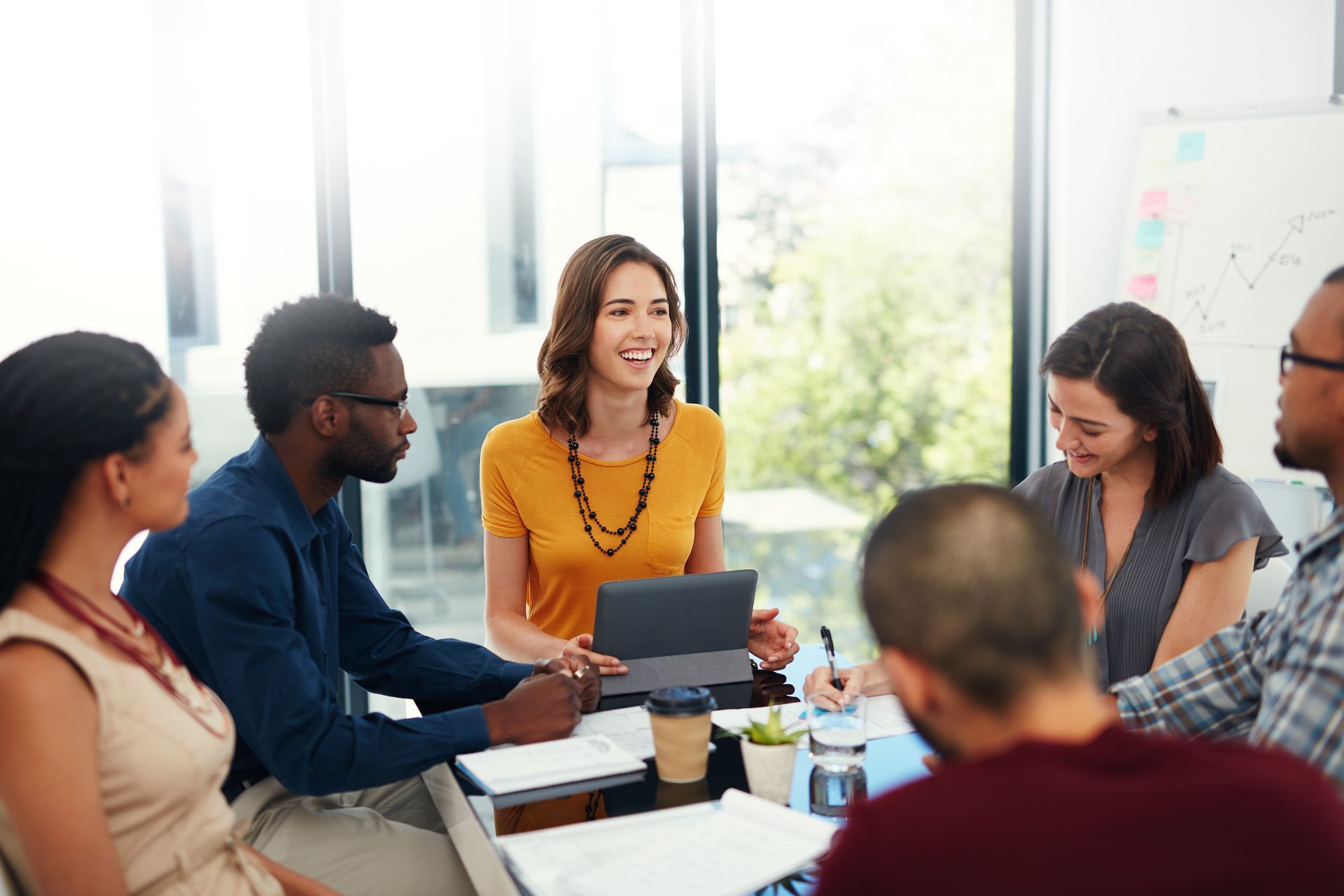 Group of diverse people in a meeting around a table, laughing. Bright room with window.