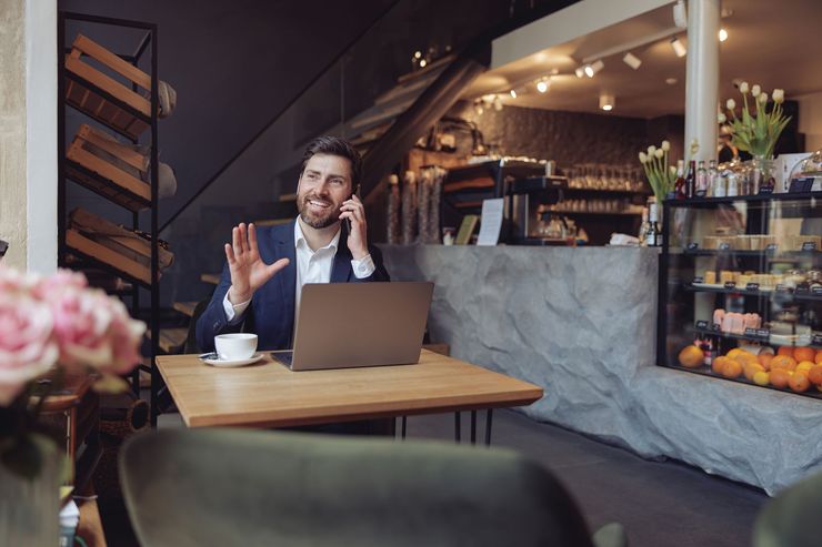 Man in blue suit at cafe, on phone, gesturing with laptop and coffee cup on table.
