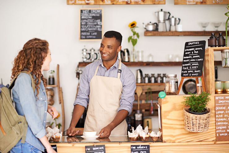 A barista smiles, handing a customer a coffee cup at a cafe counter.