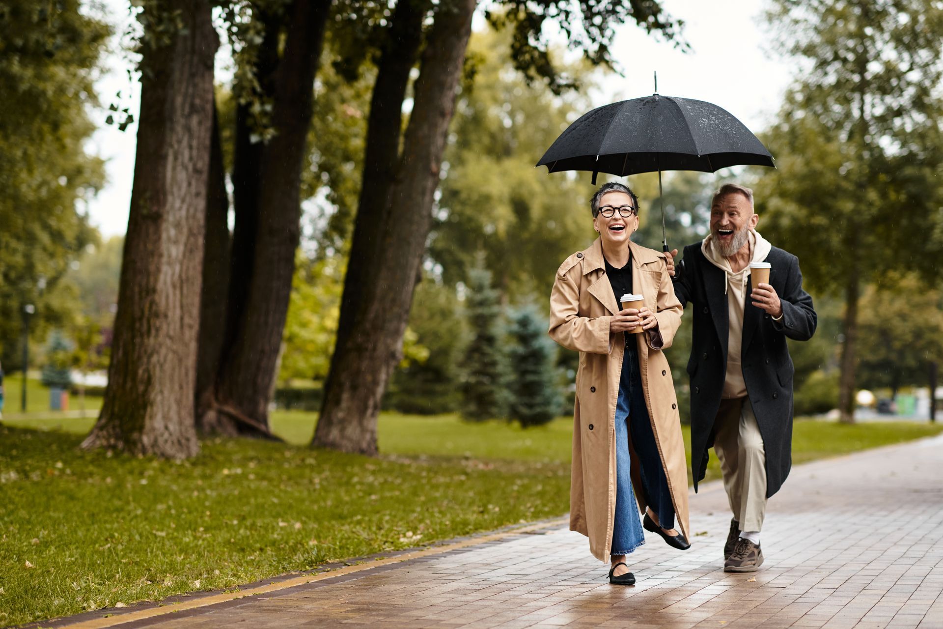 Couple under an umbrella walking and smiling in a park, holding coffee cups.
