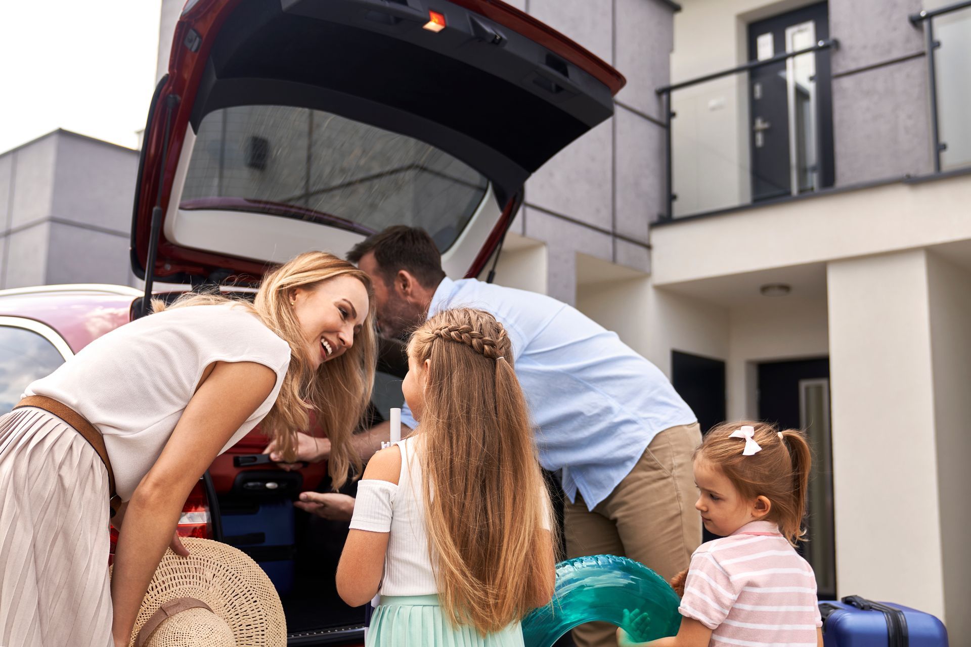 Family loading luggage into a red car trunk outside a modern house; children and parents packing.