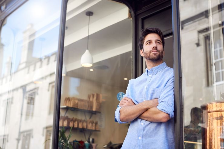 Man with crossed arms leaning in doorway, gazing outward; store interior reflected in glass.
