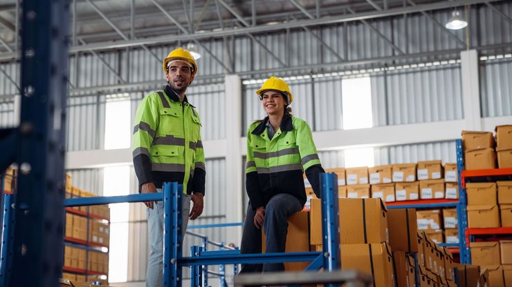 Two workers in safety vests and hard hats in a warehouse, looking towards the camera.