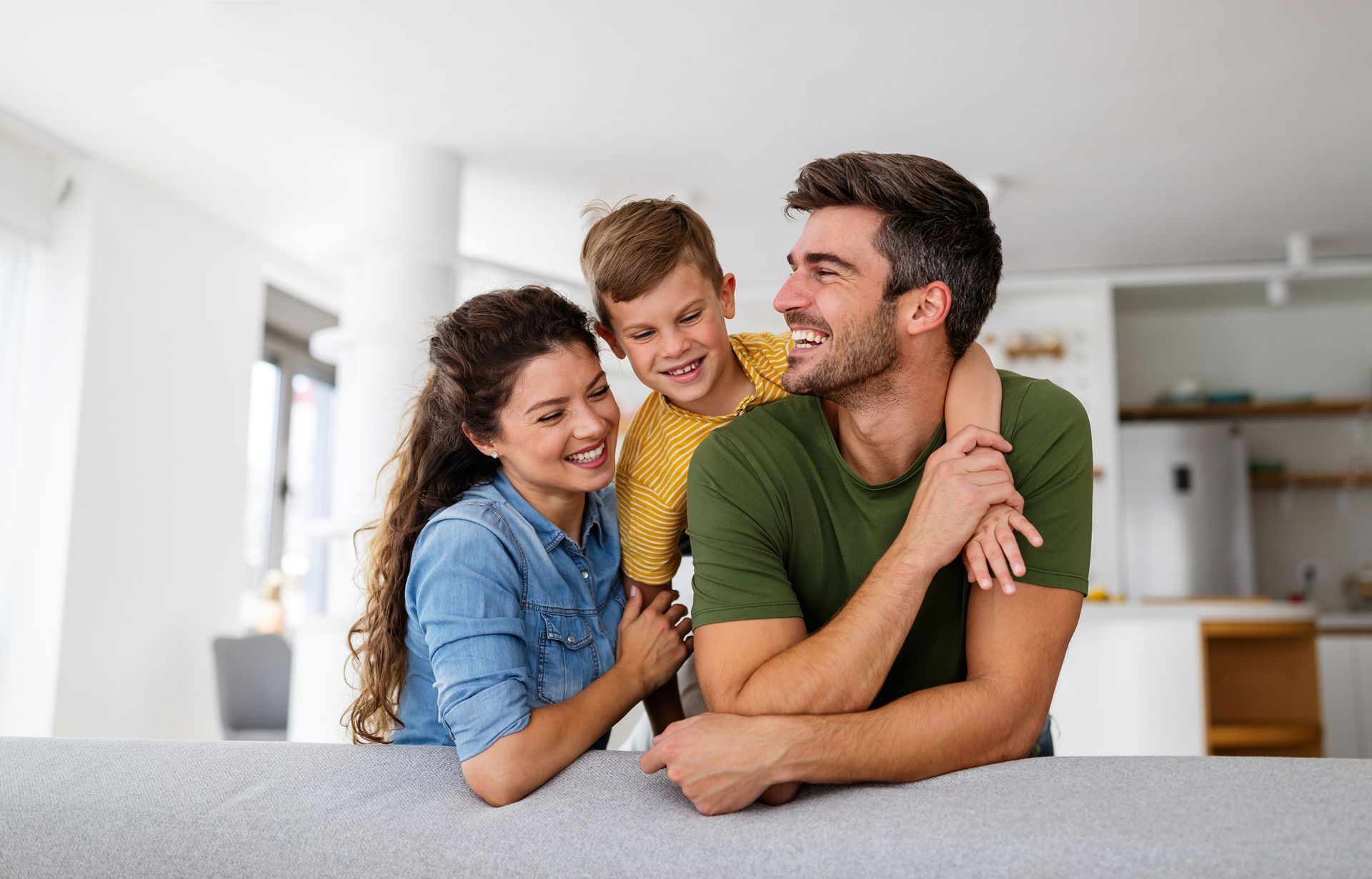 Family of three smiling, embracing on a couch in a bright living room.
