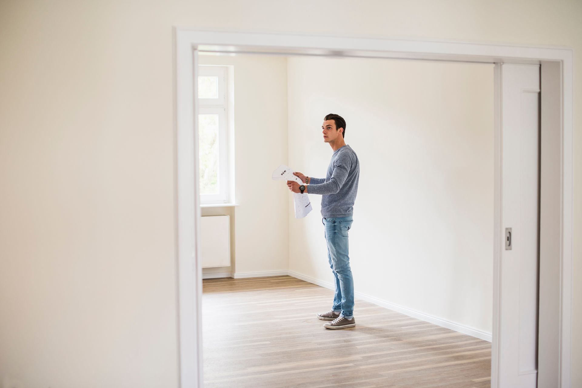 Man holding papers, standing in an empty room with a window and doorway.