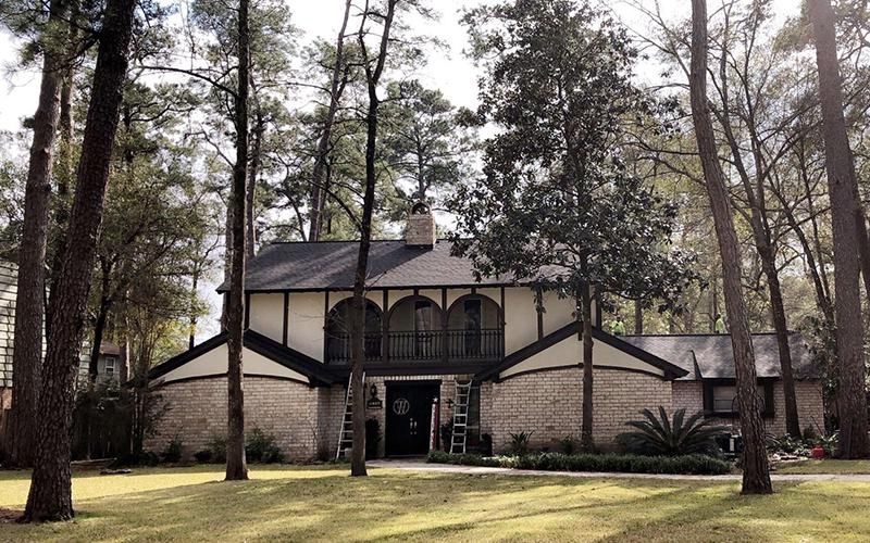 Two-story house with brick and tan facade, dark roof, framed by tall trees in a grassy yard.