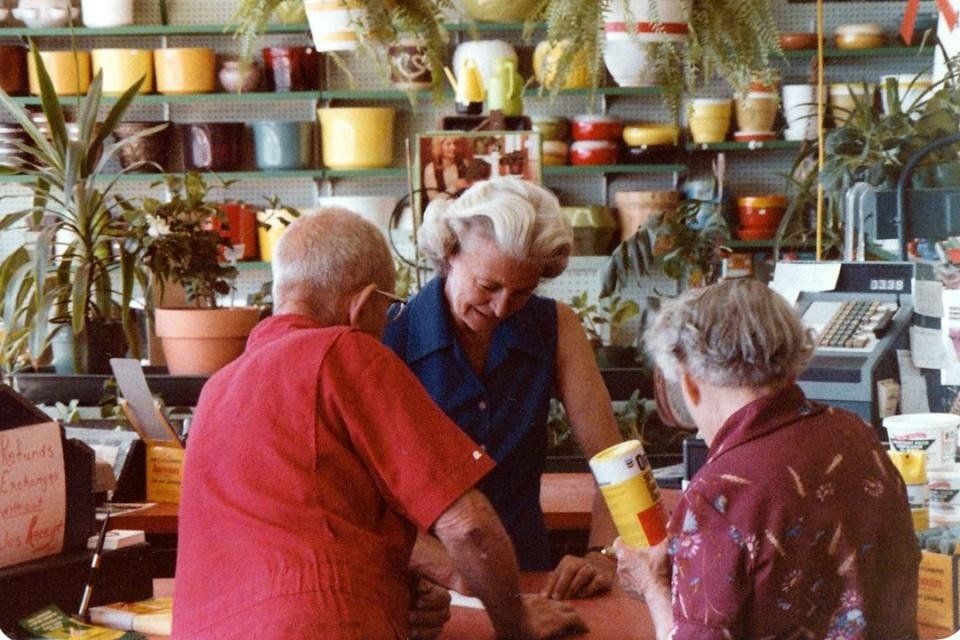 Woman assisting two customers at a plant shop counter, surrounded by plants and pots.