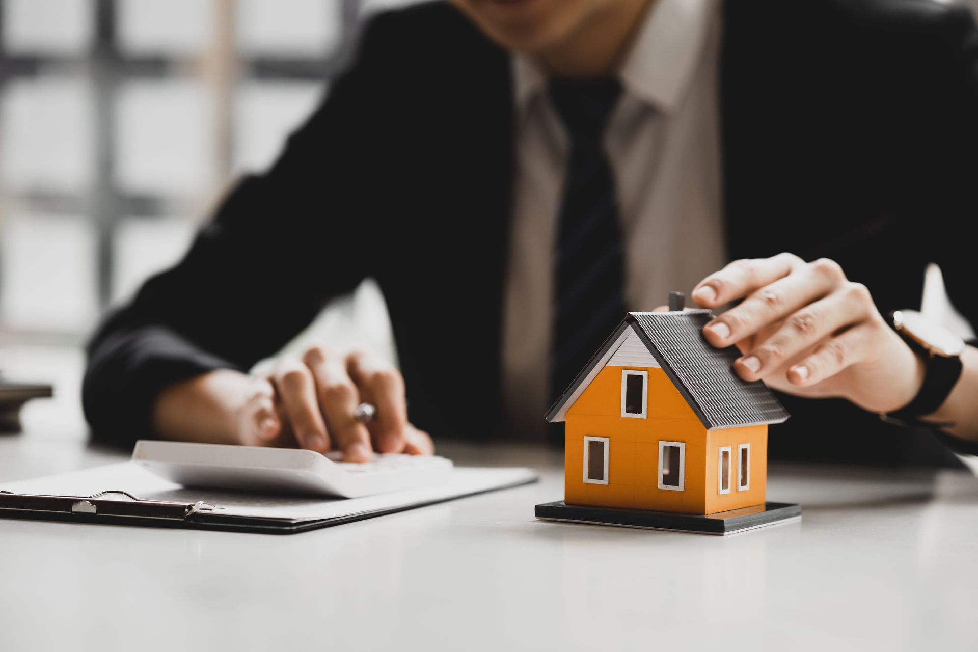 Person signs paperwork beside a small orange house model, suggesting a real estate transaction.