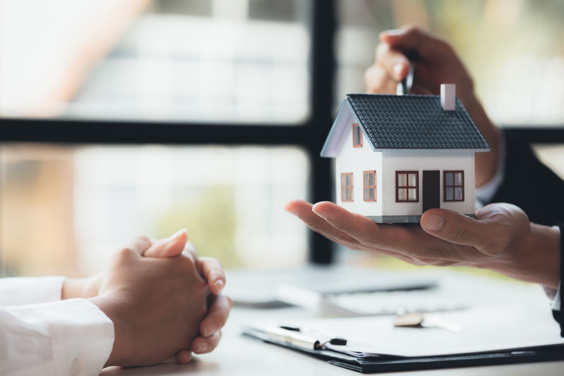 Hands holding a model house; a pen is offered. A person sits at a desk, possibly buying or selling property.