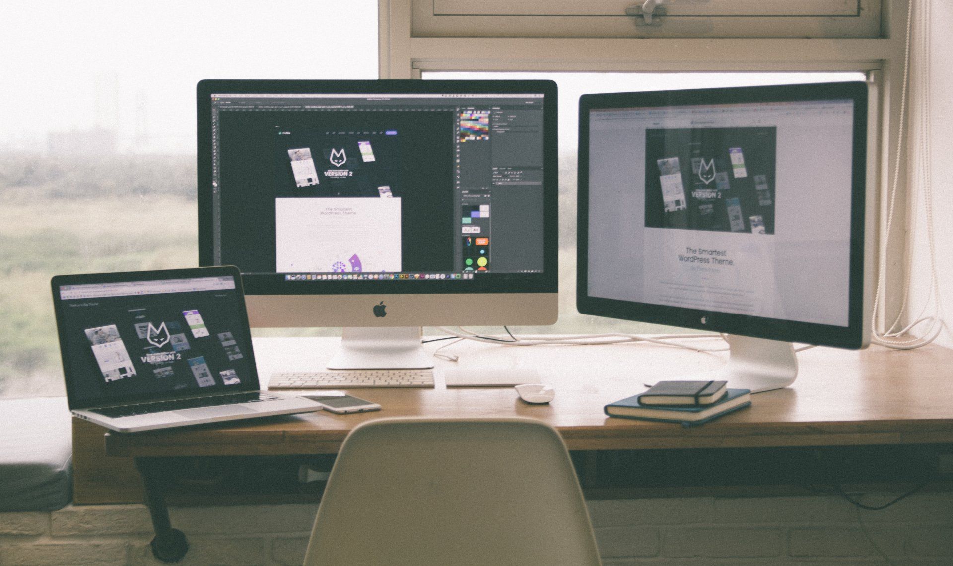 Desk with laptop and two monitors showing design mockups in a bright office workspace