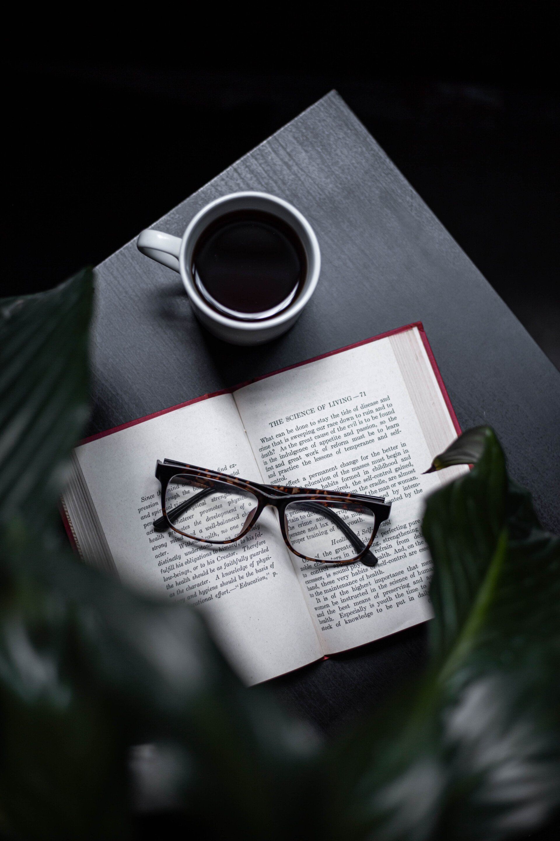 Coffee cup, open book, and eyeglasses on a dark desk with soft plant shadows.