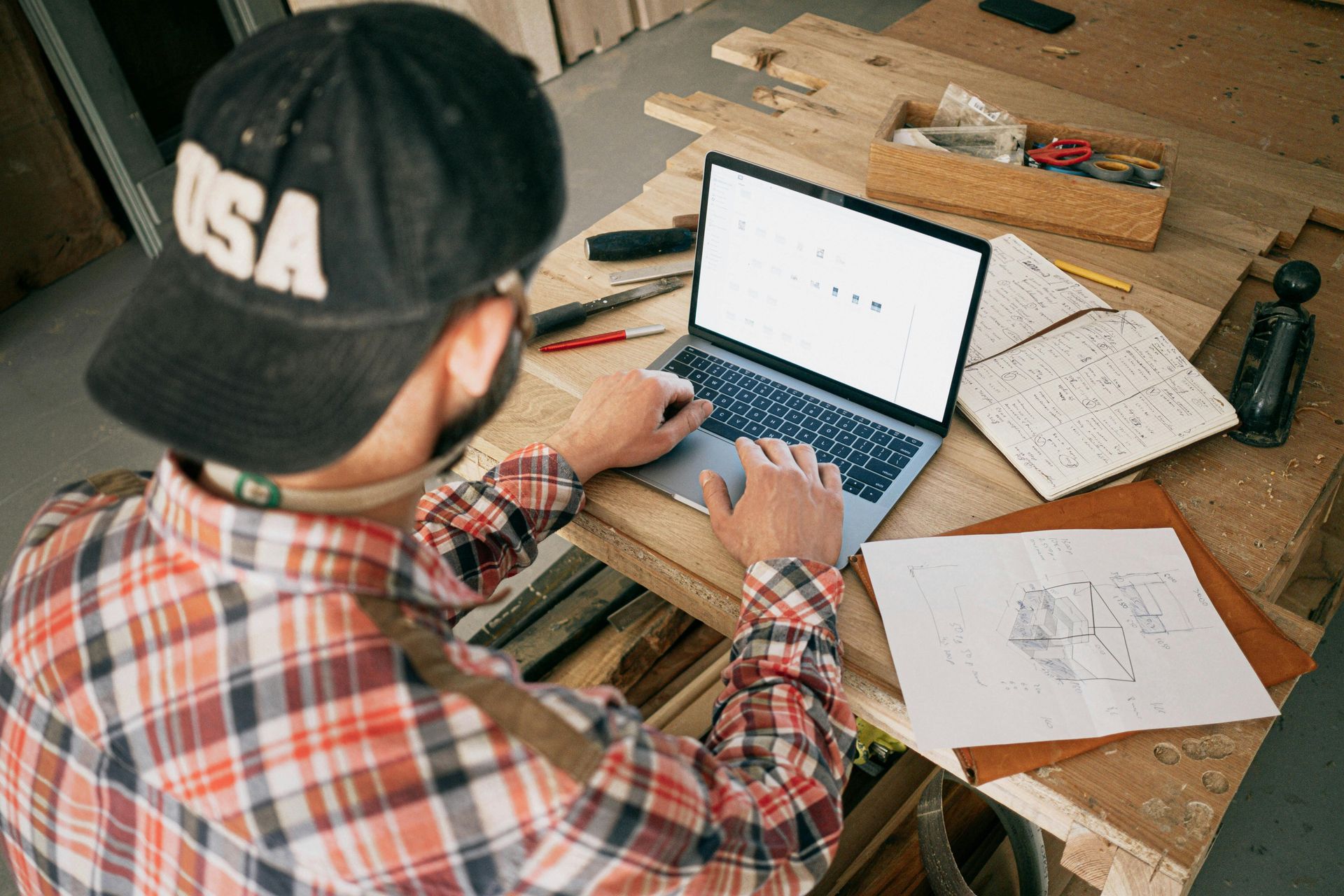 Person wearing a plaid shirt and USA cap types on a laptop at a workbench with tools and sketches.