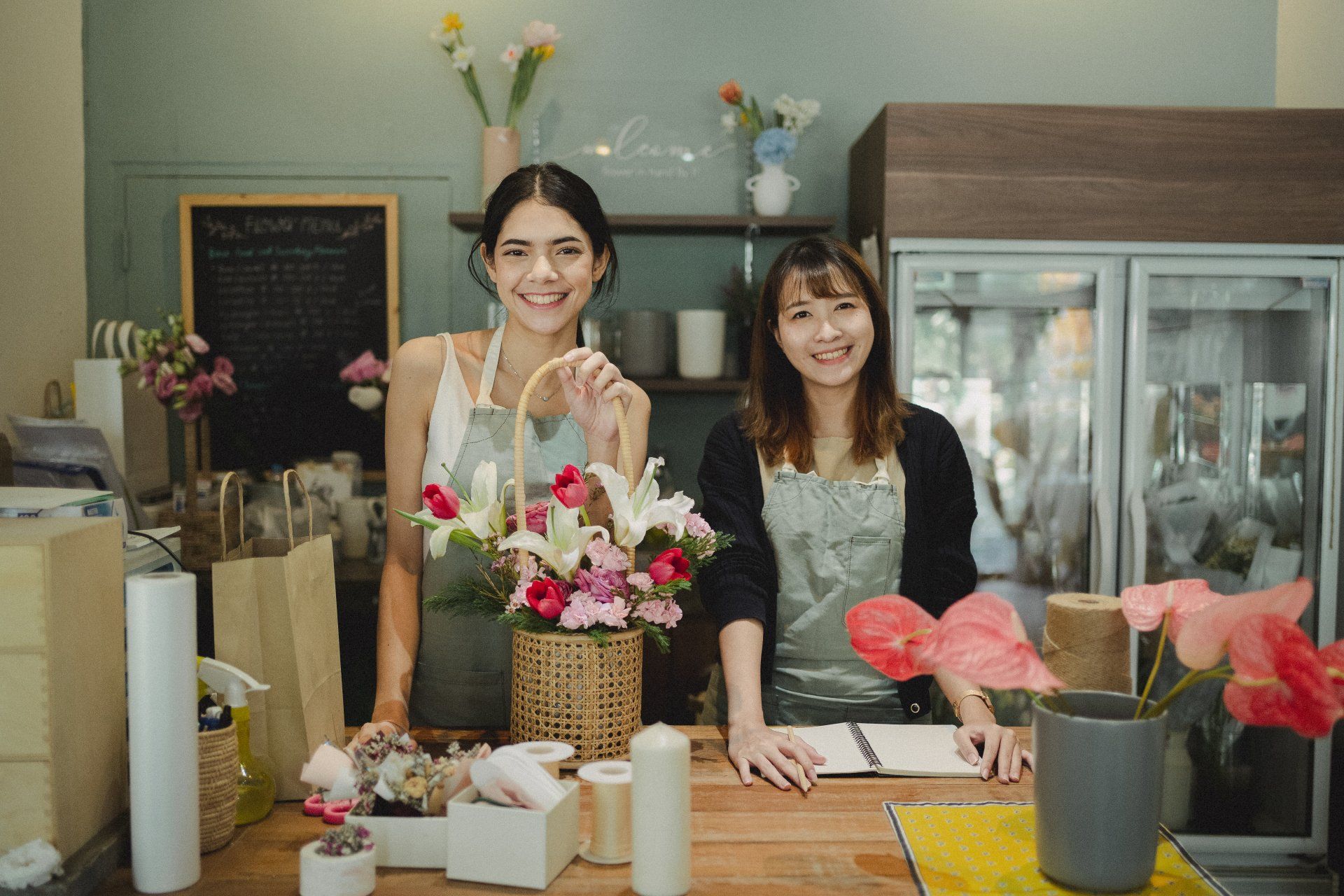 Two women arranging flowers behind a shop counter, with bouquets, vases, and a display fridge in the background