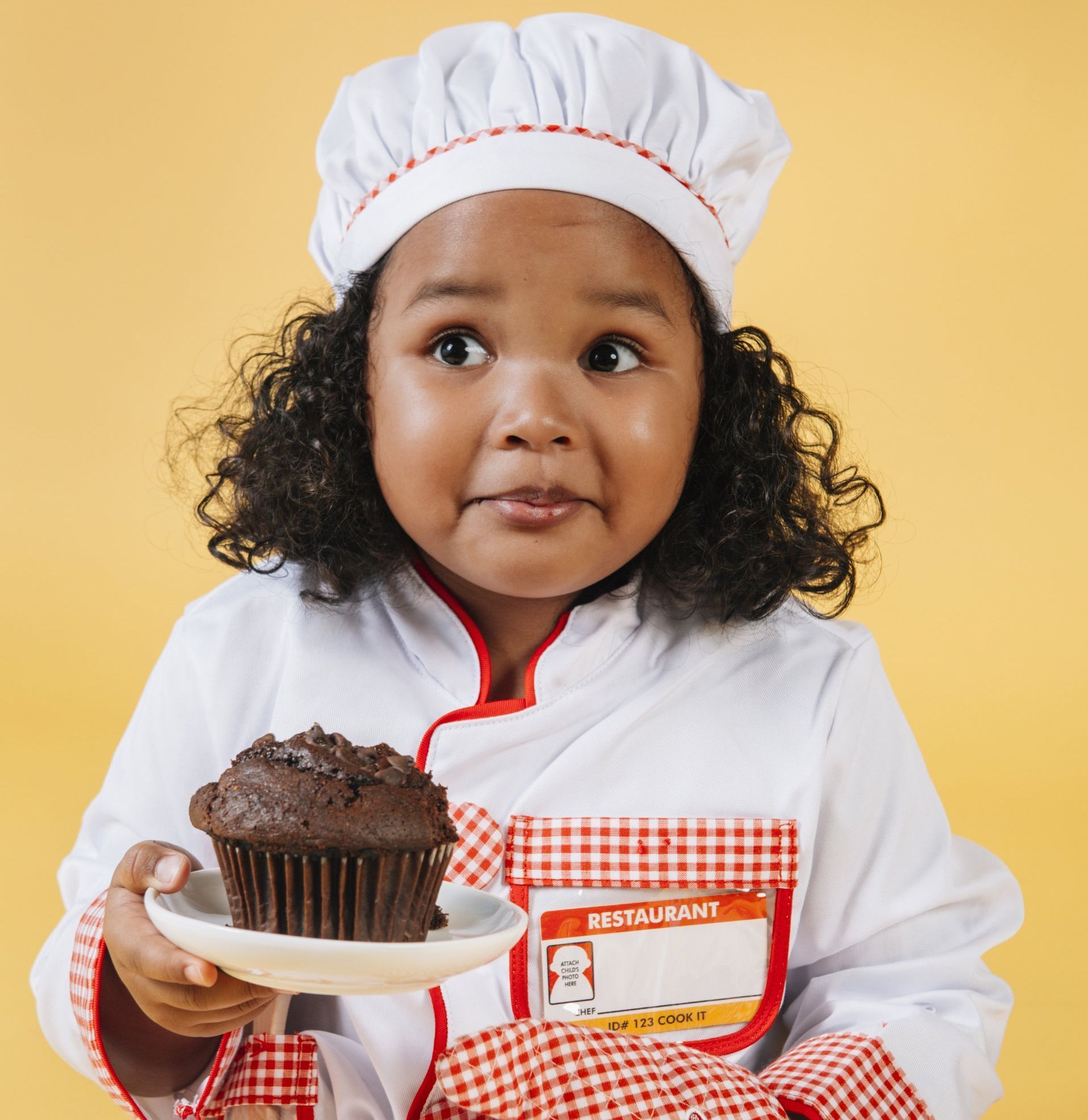 Child chef in a white uniform and hat holding a plate with a chocolate cupcake on a yellow background
