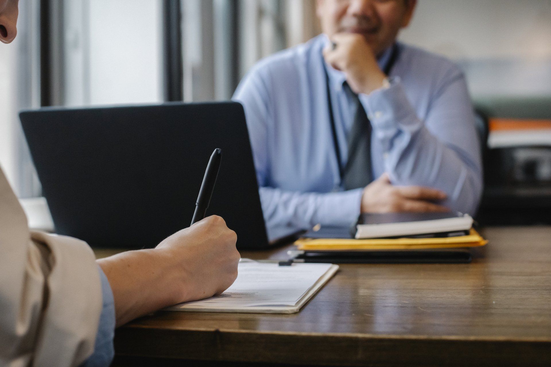 Two people in a business meeting at a table with a laptop and notebook, one writing while the other listens.