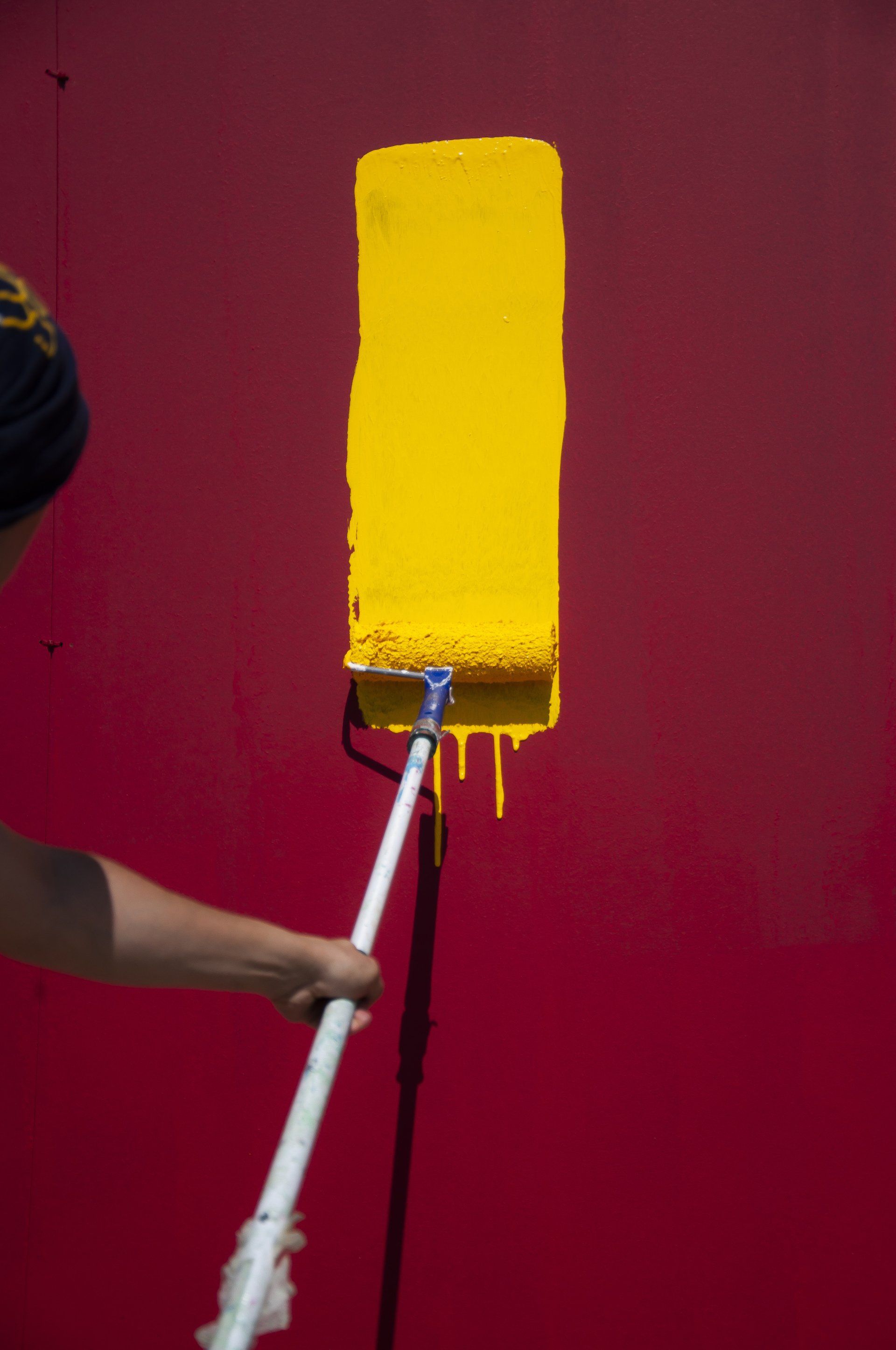 Person painting a maroon wall with a bright yellow roller in a narrow vertical section.