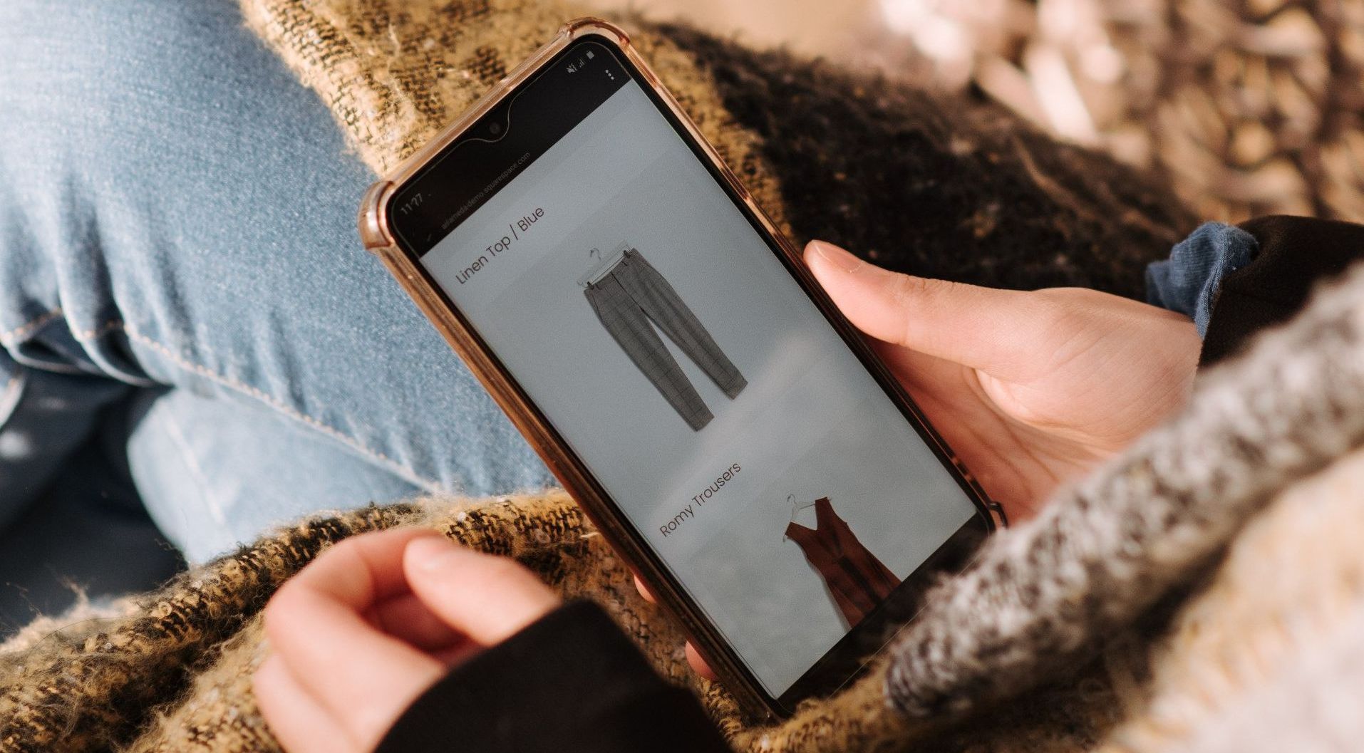 Person using a smartphone to browse clothing while sitting on a couch with a blanket