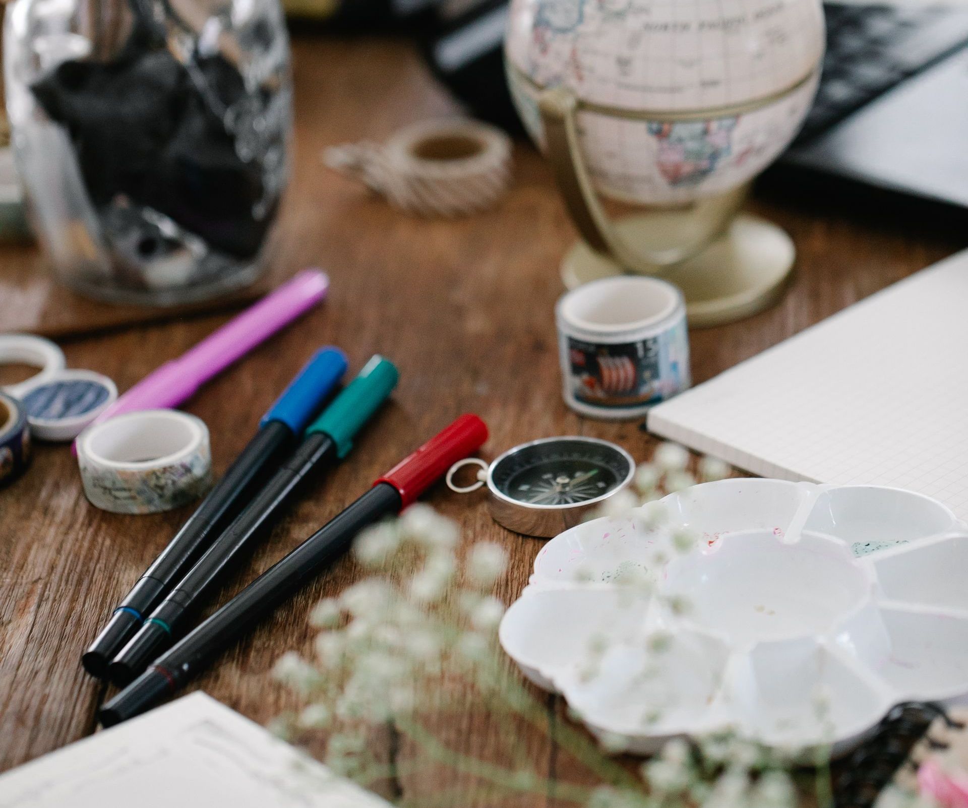 Art supplies on a wooden desk: pens, washi tape, a globe, and a white paint palette