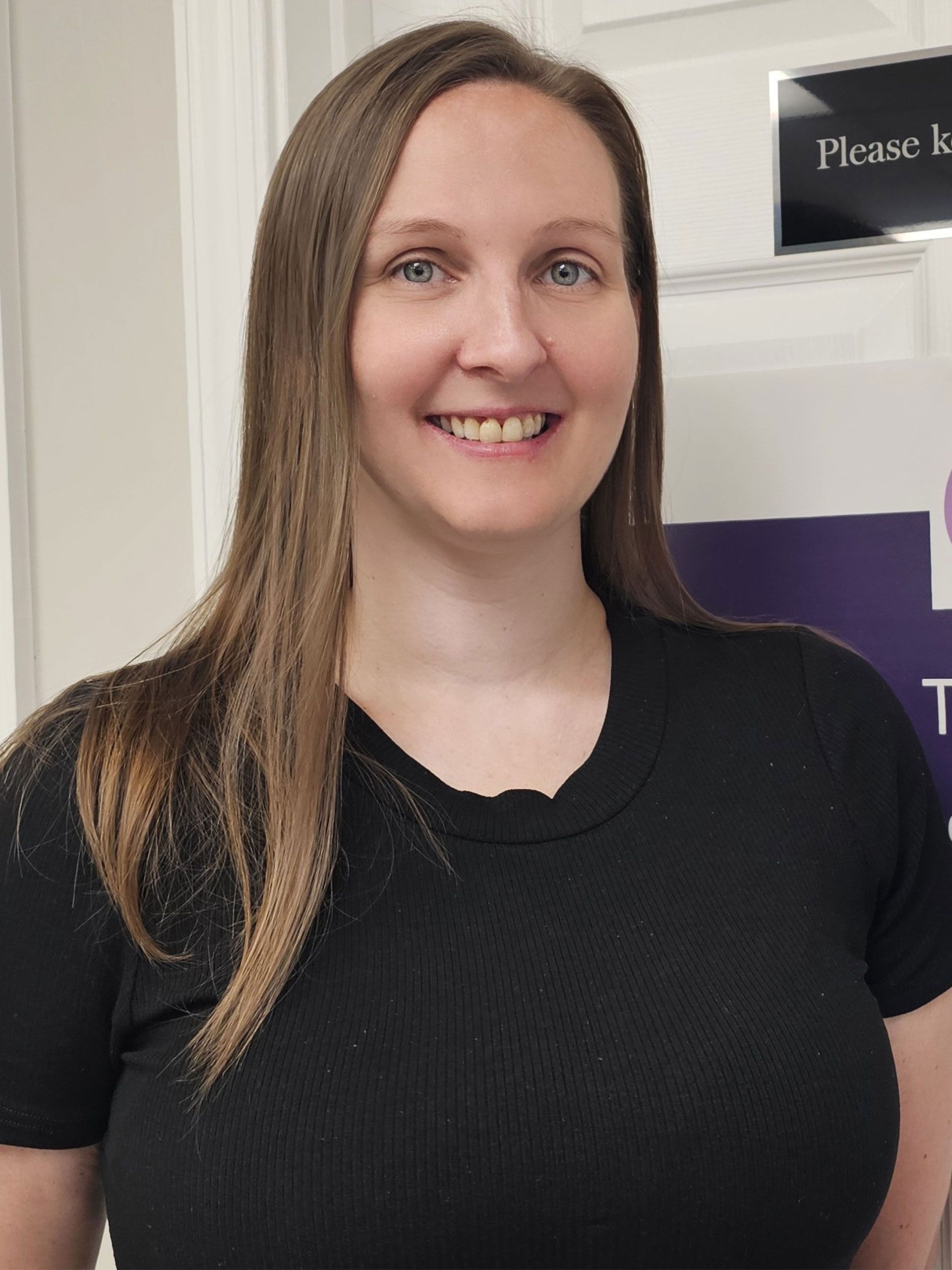 Woman smiling indoors in a black shirt, standing beside a purple sign.