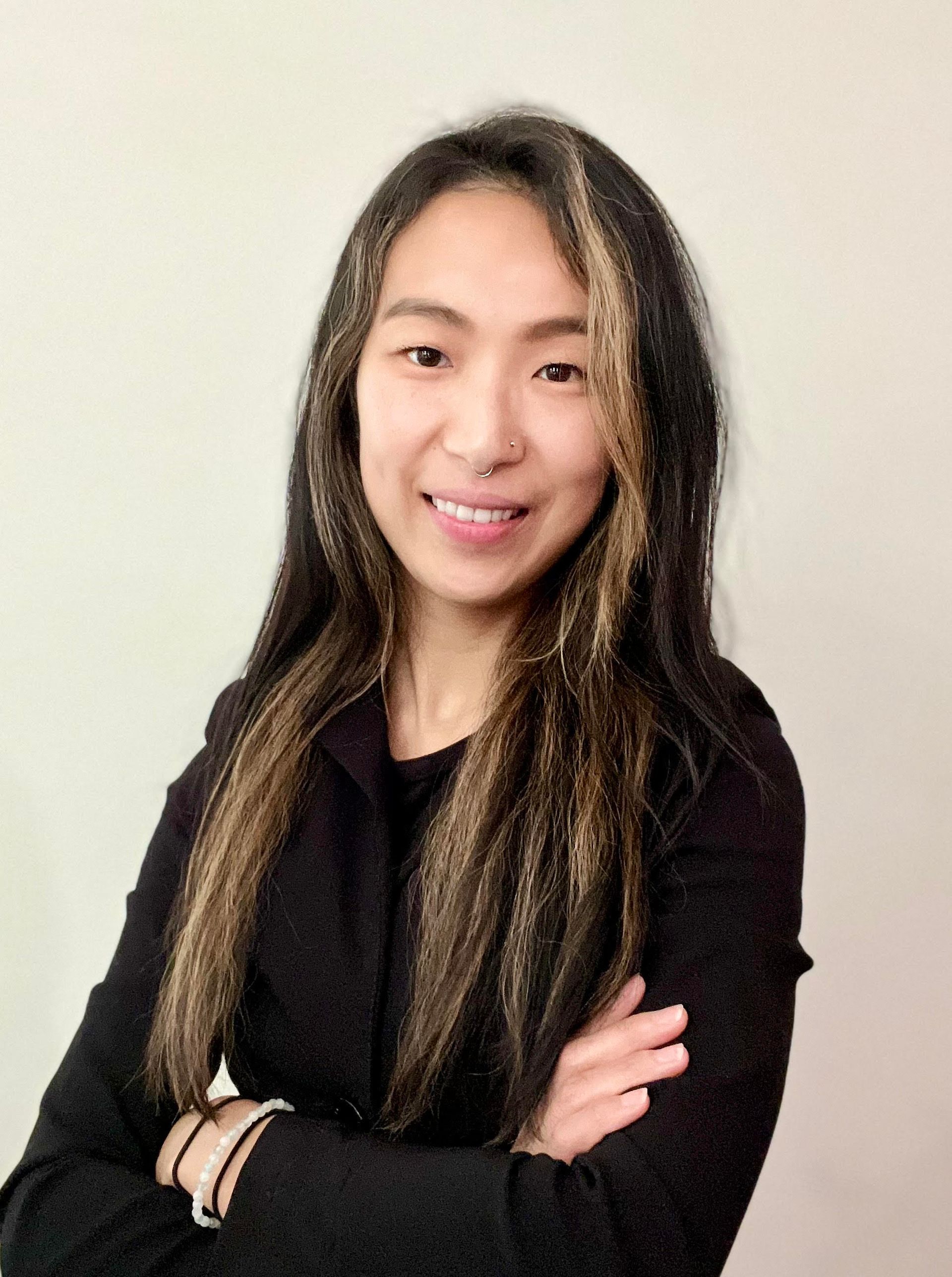 Smiling person with long hair and black top, standing with arms crossed against a plain light background