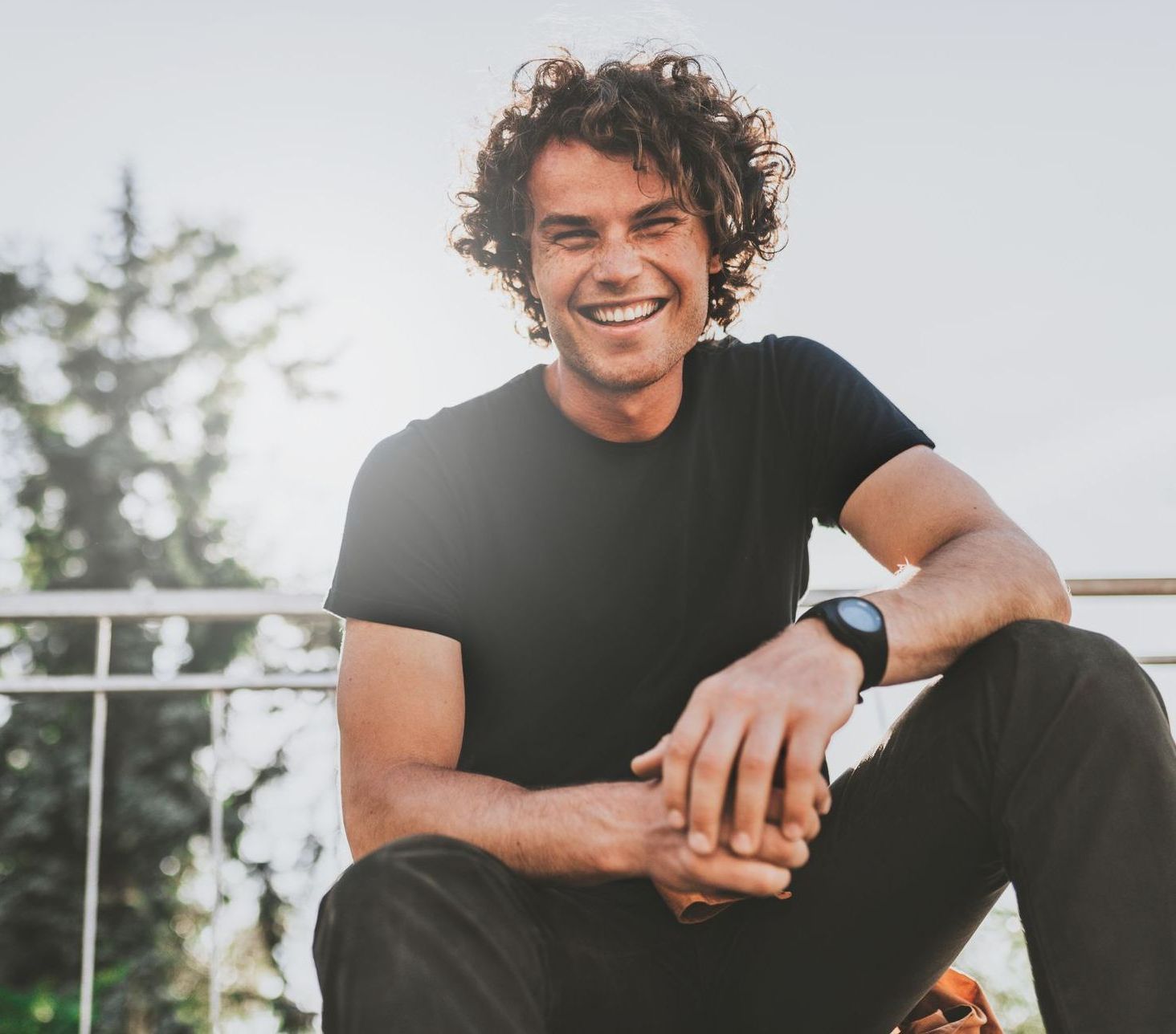 Smiling man in black shirt sitting outdoors on a railing, with trees in the background