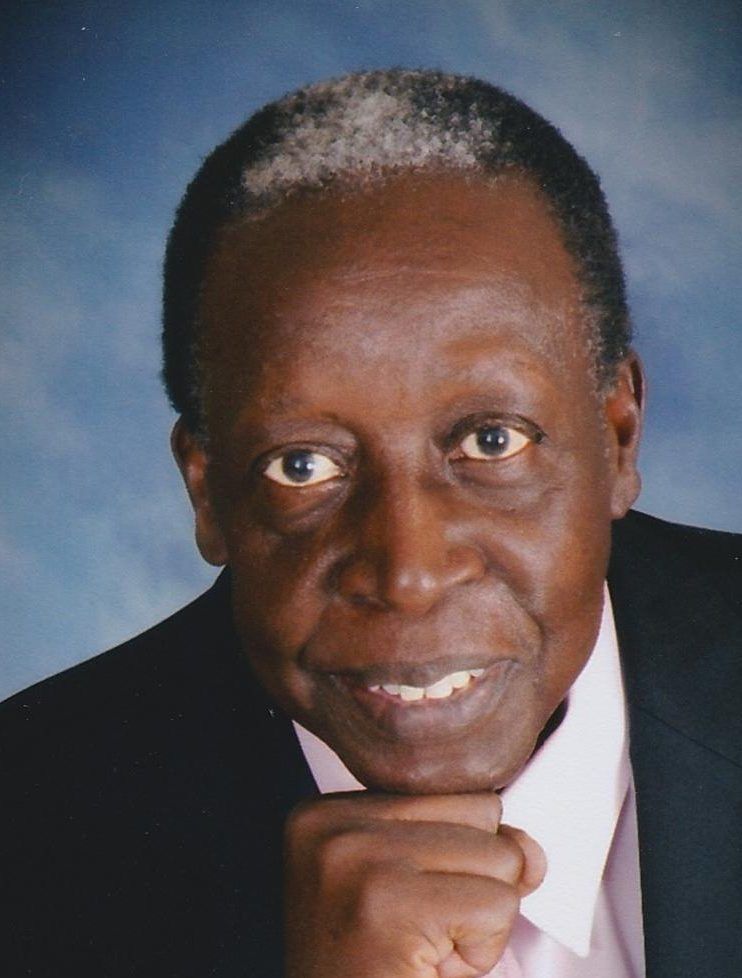 A man in a suit and pink shirt is smiling with his hand on his chin.