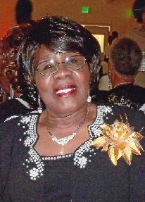 A woman wearing glasses and a flower pin is smiling for the camera.