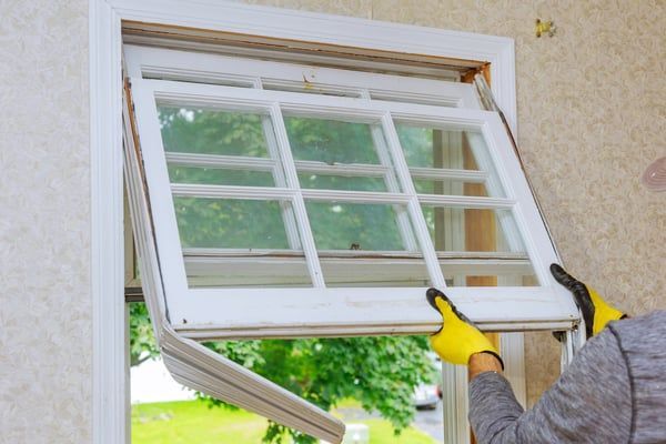 Person installing a white-framed window, lifting it into a wall opening with yellow gloves