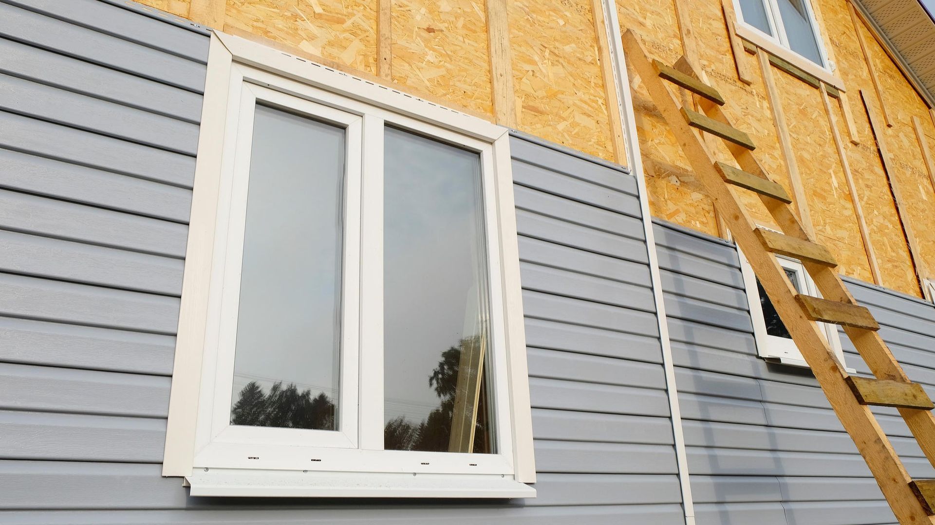 White window in gray siding next to exposed plywood wall and ladder during home construction