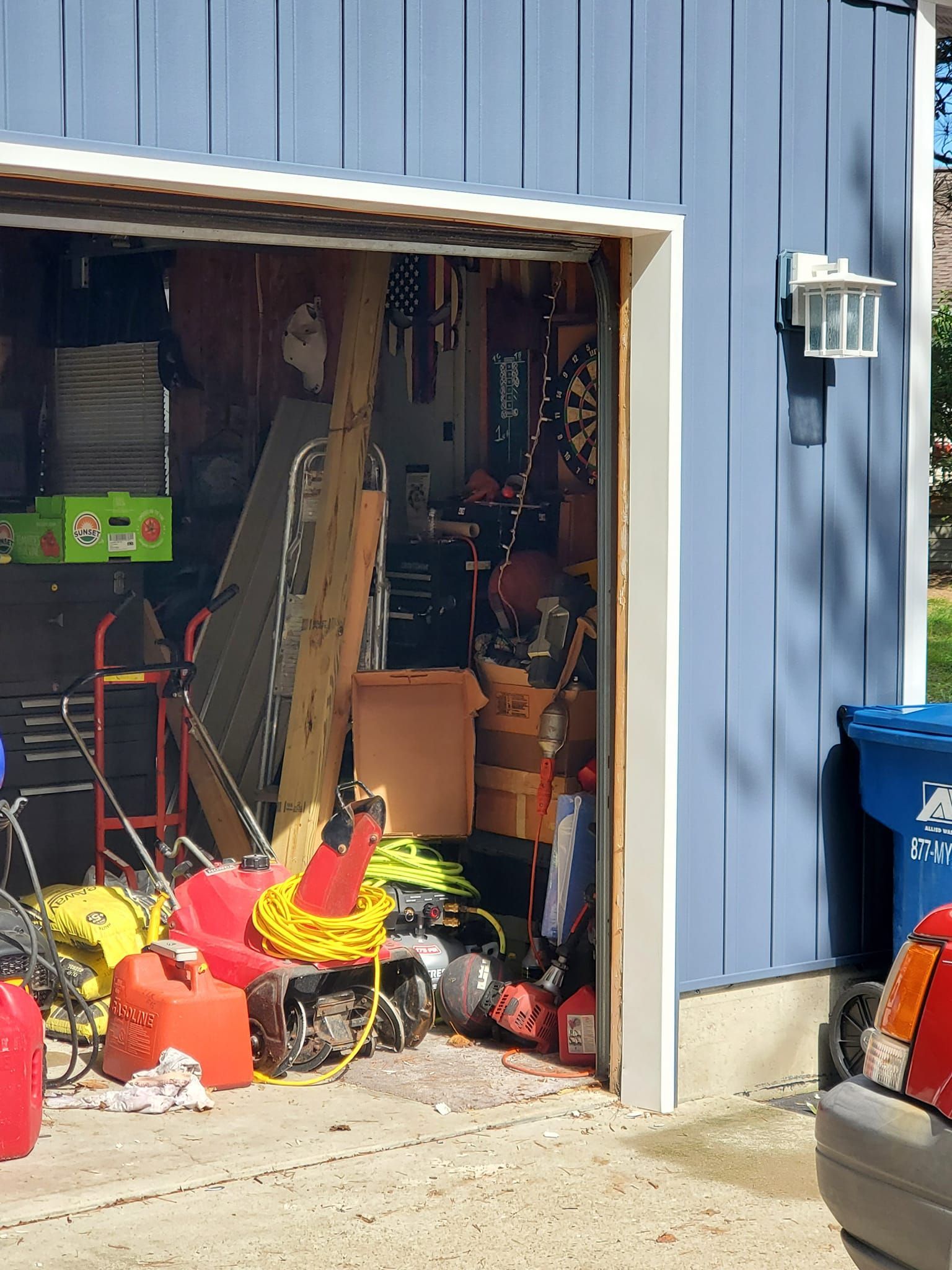 Open garage with cluttered tools, red equipment, and boxes beside a blue house wall