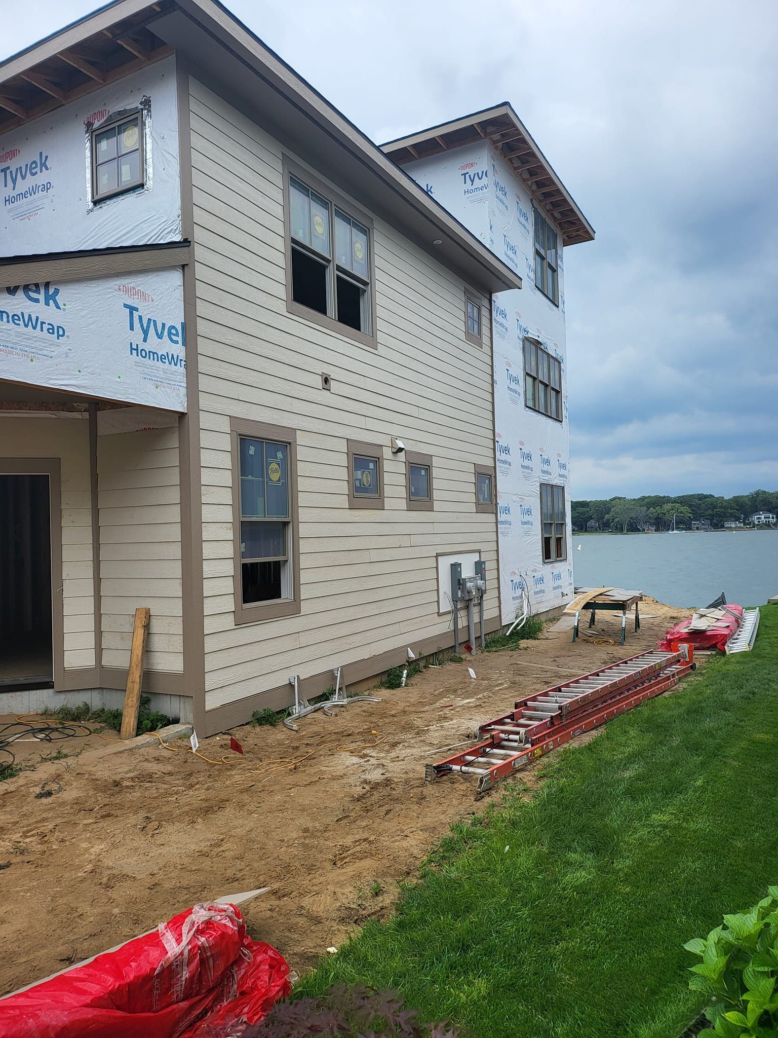 Two-story house under construction with white siding, muddy yard, and red safety netting beside green grass.