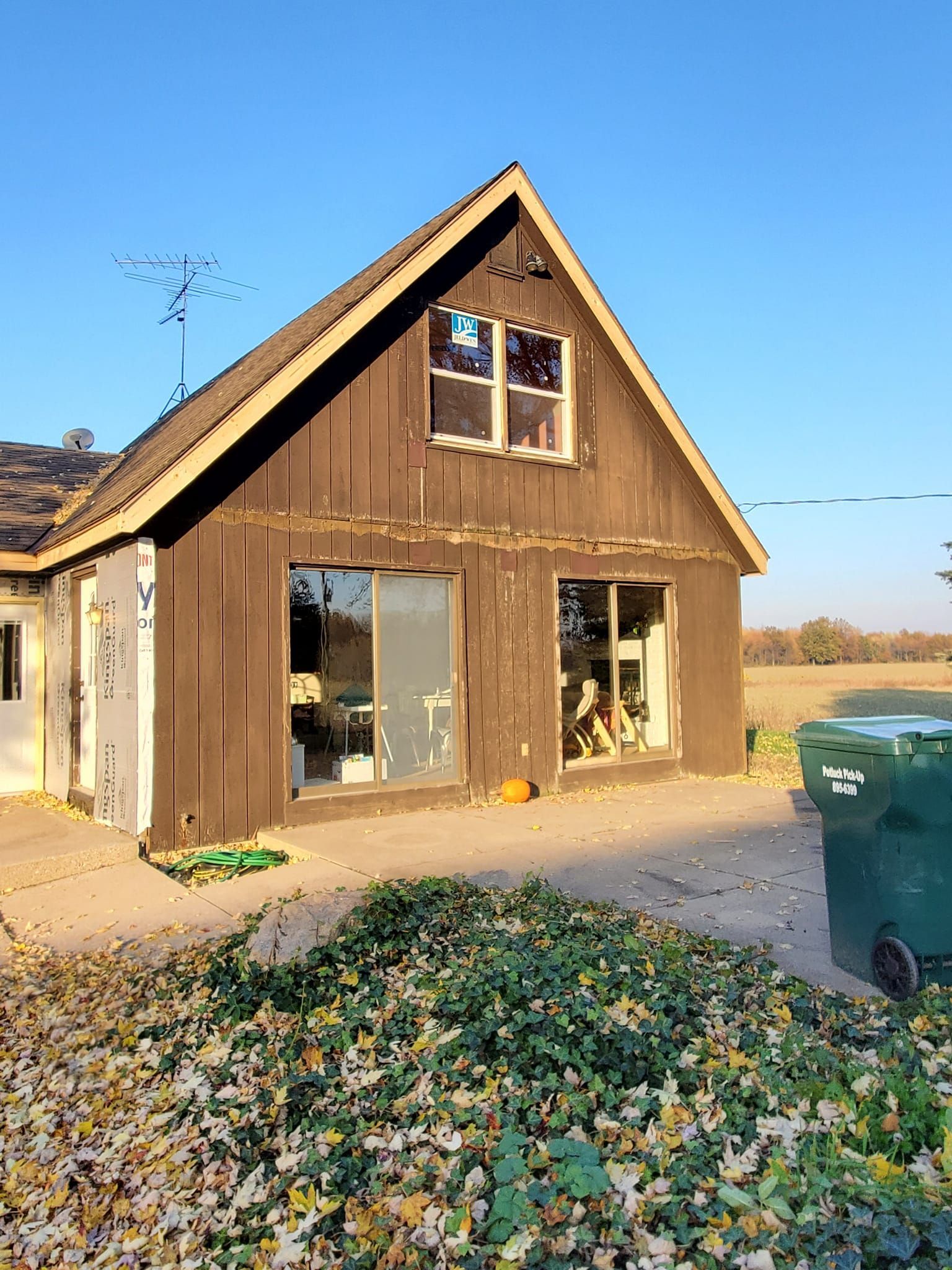 Small wooden house under construction with a large leaf pile in front and a green trash bin at right.