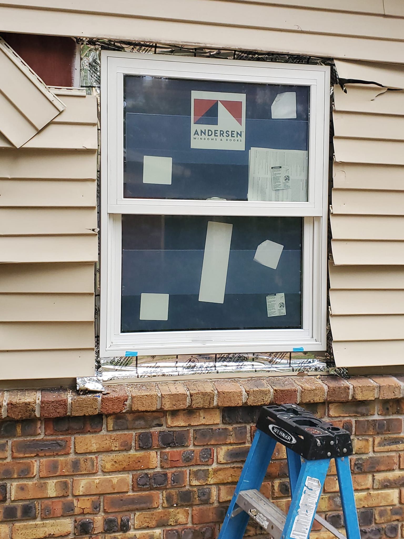 Window with blue paper coverings and a ladder leaning against a brick-and-siding house exterior.