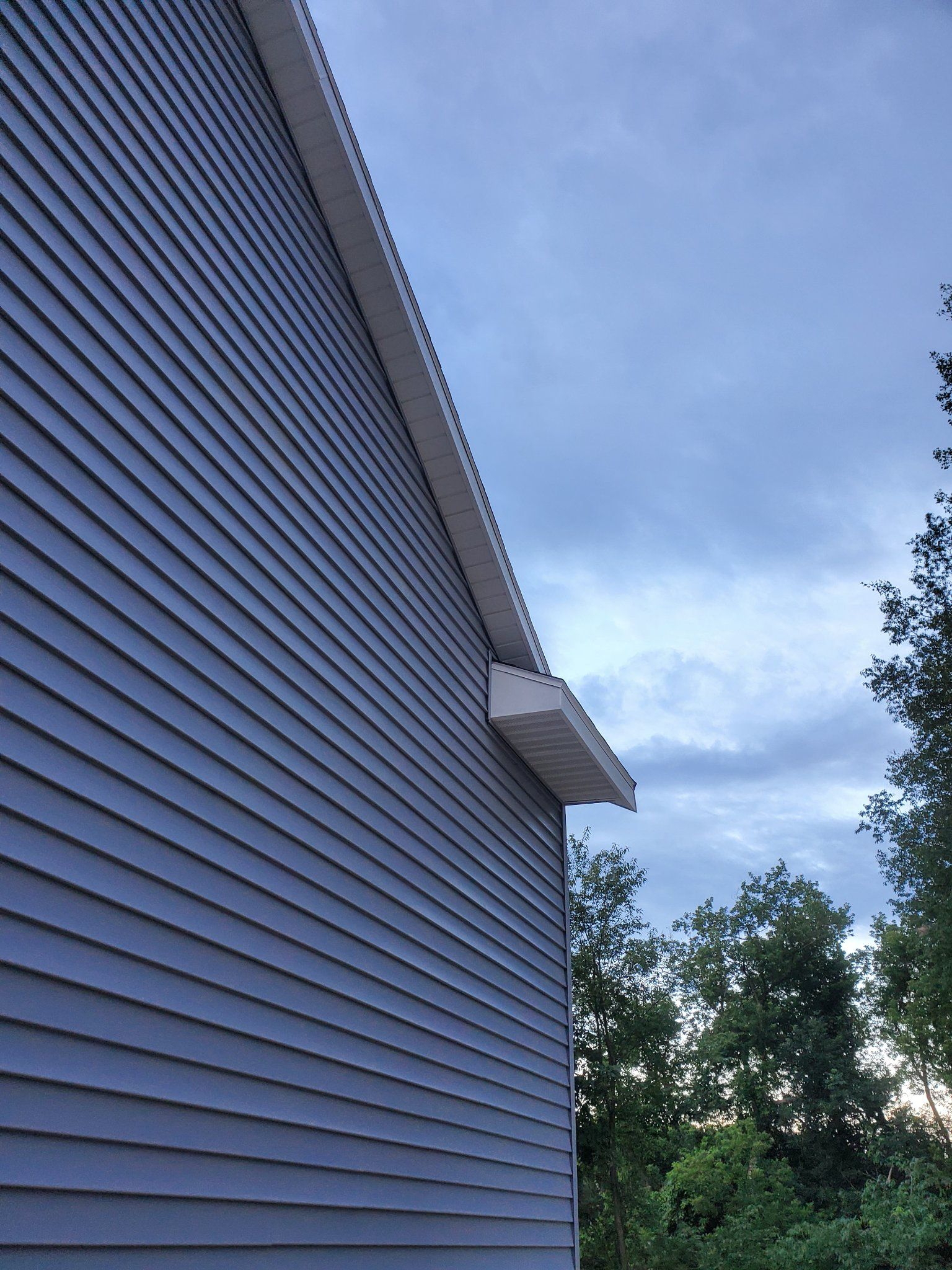 Blue-sided house exterior with a white vent and trees under a cloudy sky