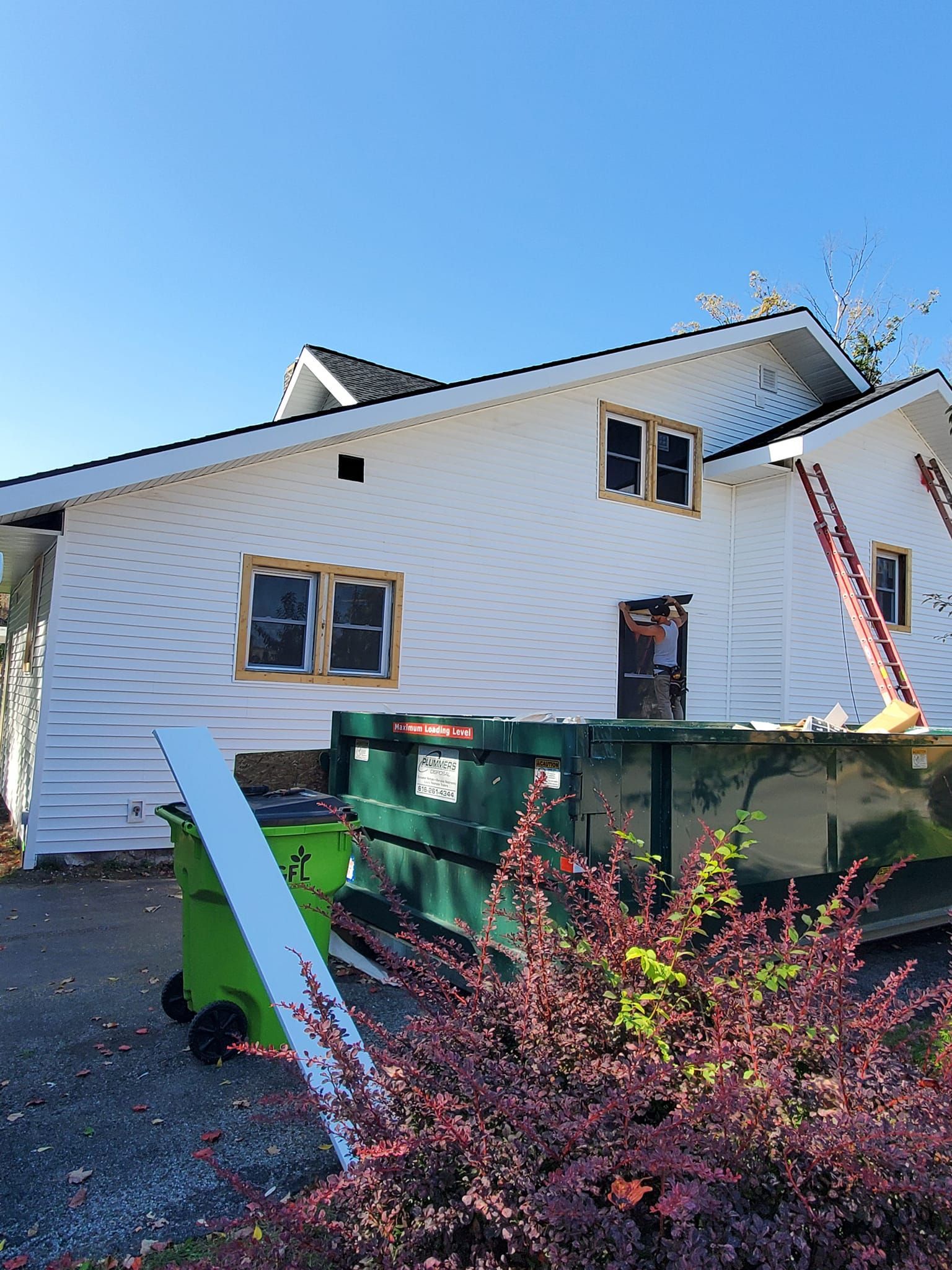 Damaged white house with broken windows beside a green dumpster and pink bushes under a blue sky