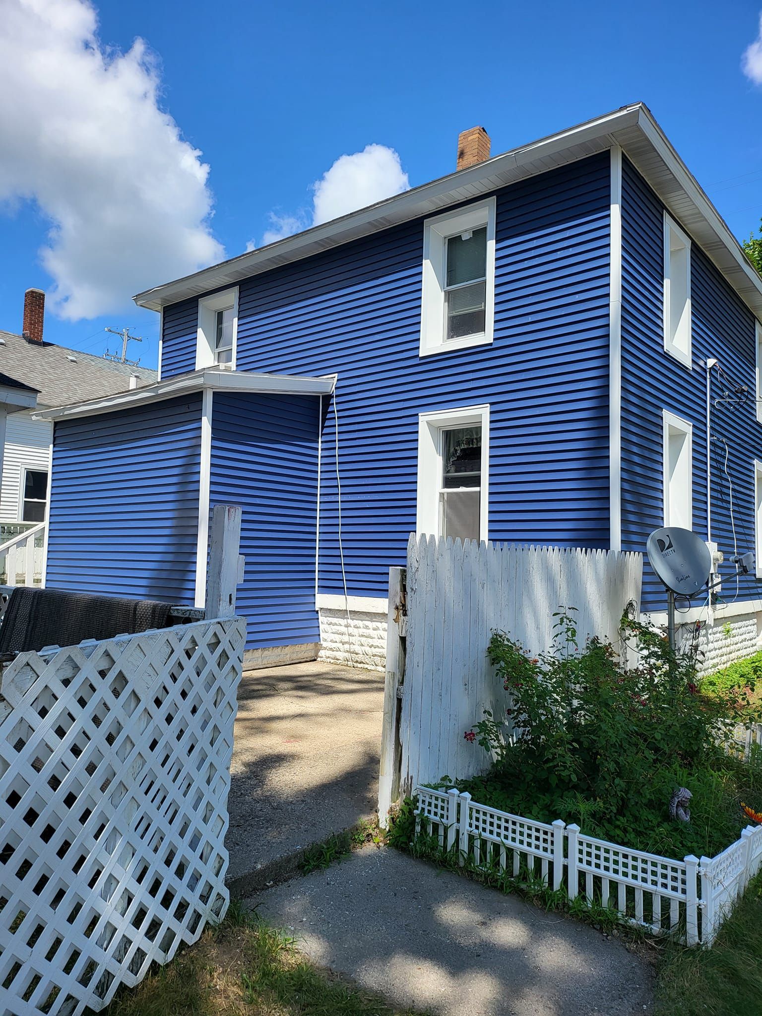 Blue two-story house with white trim, a small front path, and a white lattice fence under a partly cloudy sky