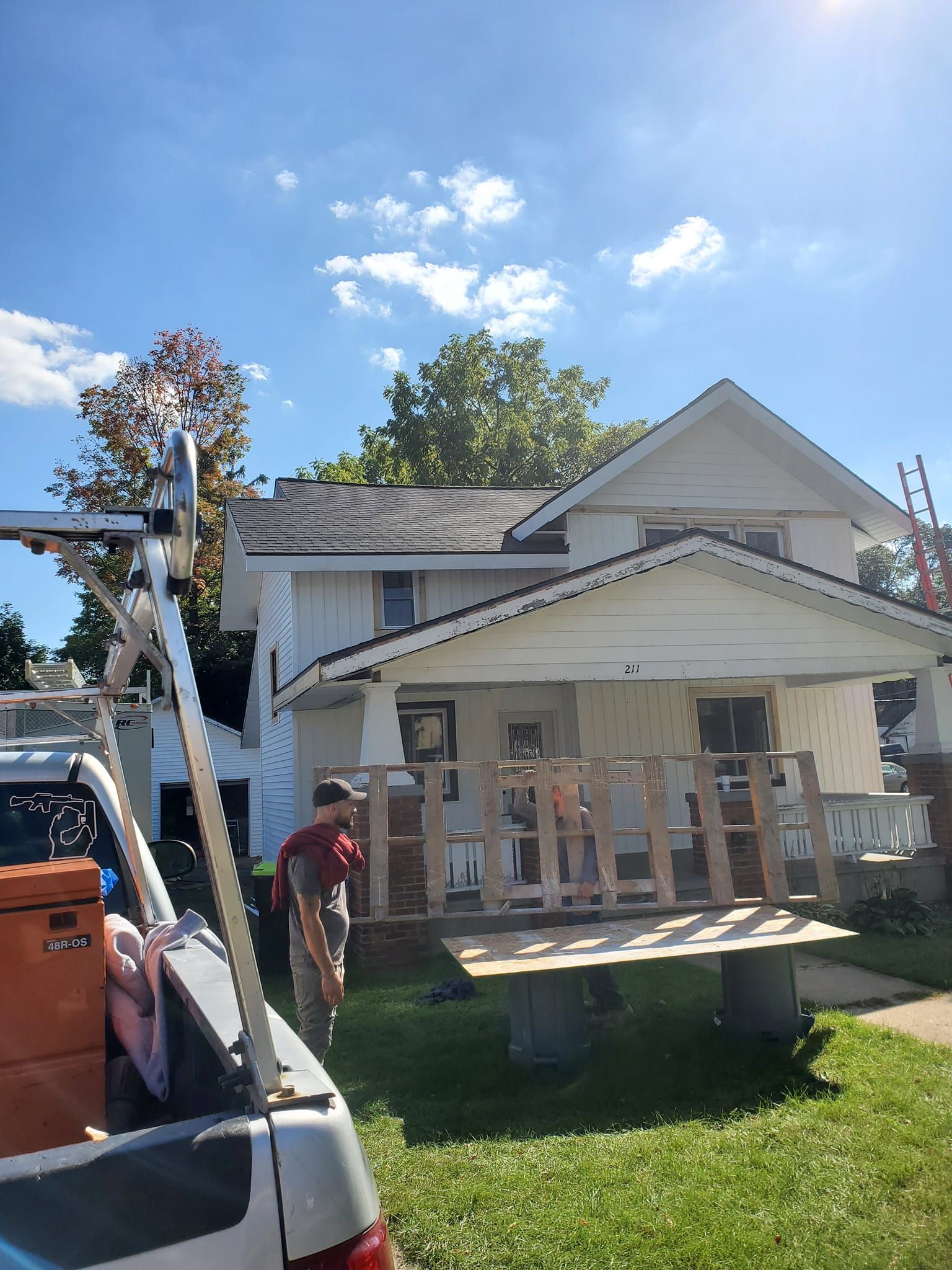 House with porch under construction, with ladder, tools, and workers outside on a sunny day