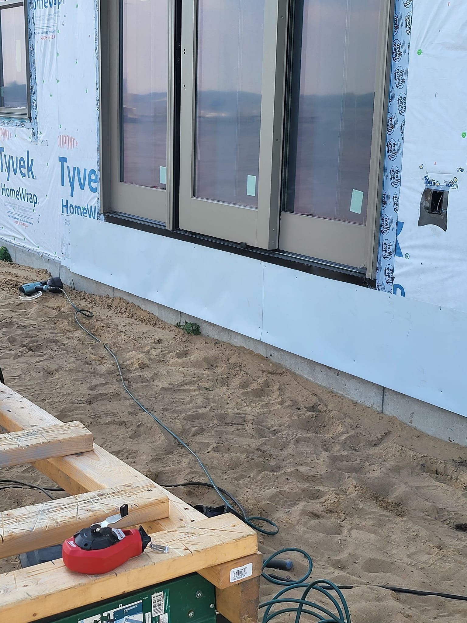 House exterior under construction with unfinished siding, windows, dirt ground, and a red tool on a wooden frame.