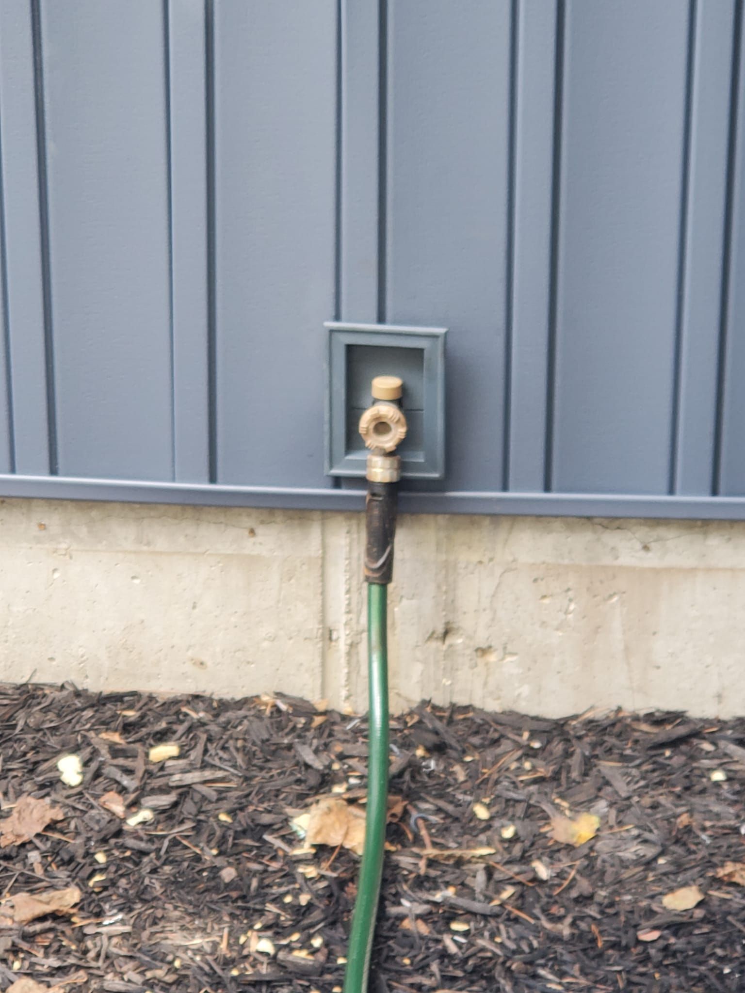 Outdoor water spigot with green hose attached against gray siding and concrete foundation